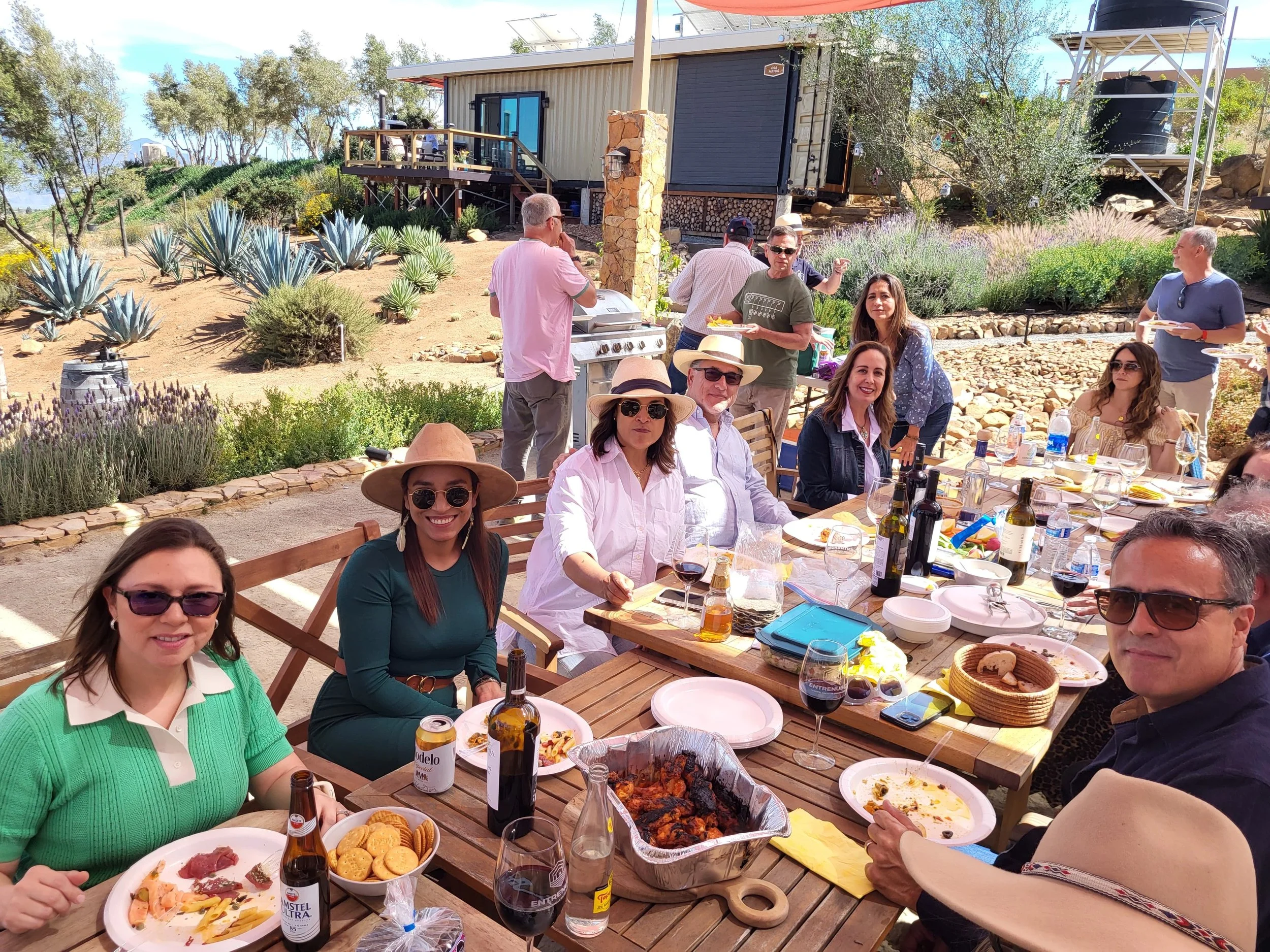 A group of people enjoying an outdoor family gathering with food and drinks at a wooden table in a scenic garden, with a house and trees in the background.