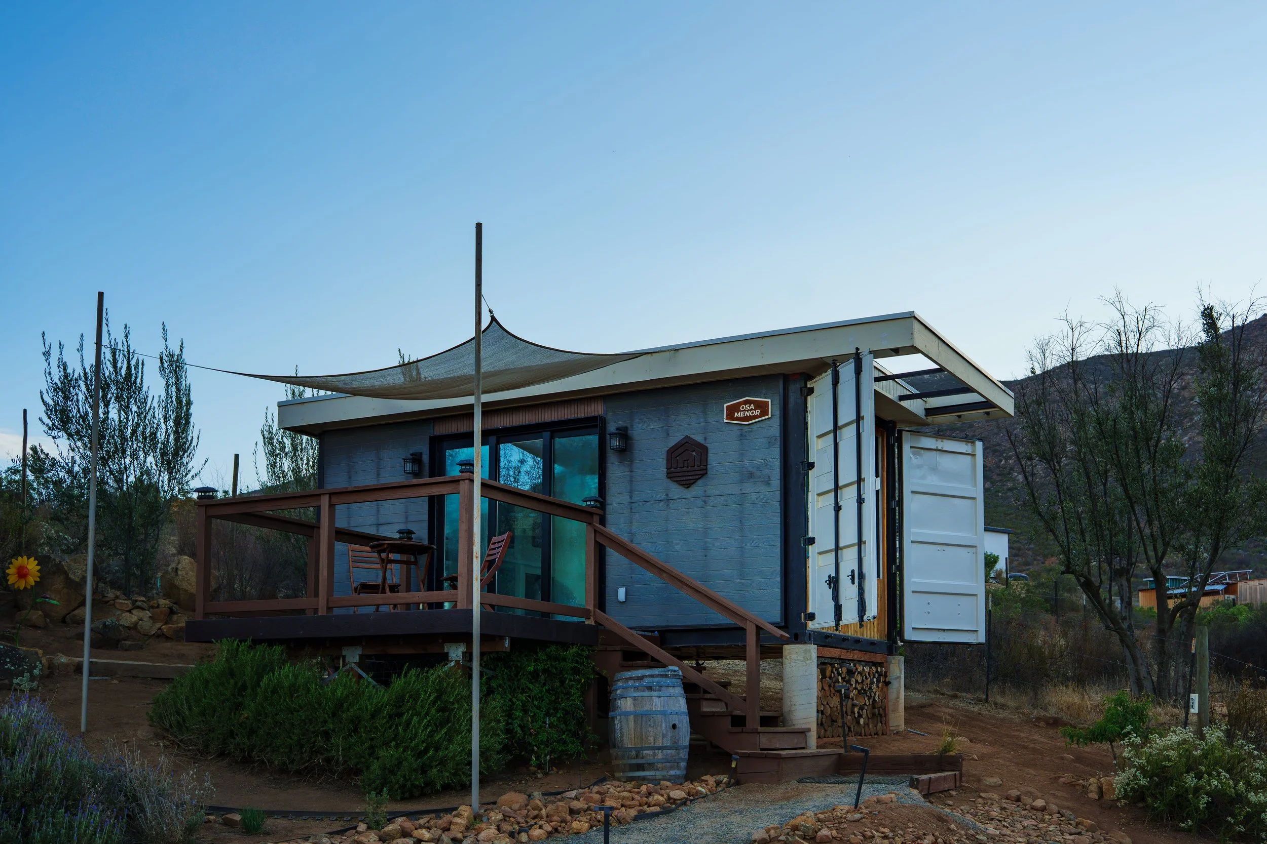 A small modern house built on a hillside with a wooden deck, glass doors, and a shade sail overhead, surrounded by greenery and trees under a clear blue sky.
