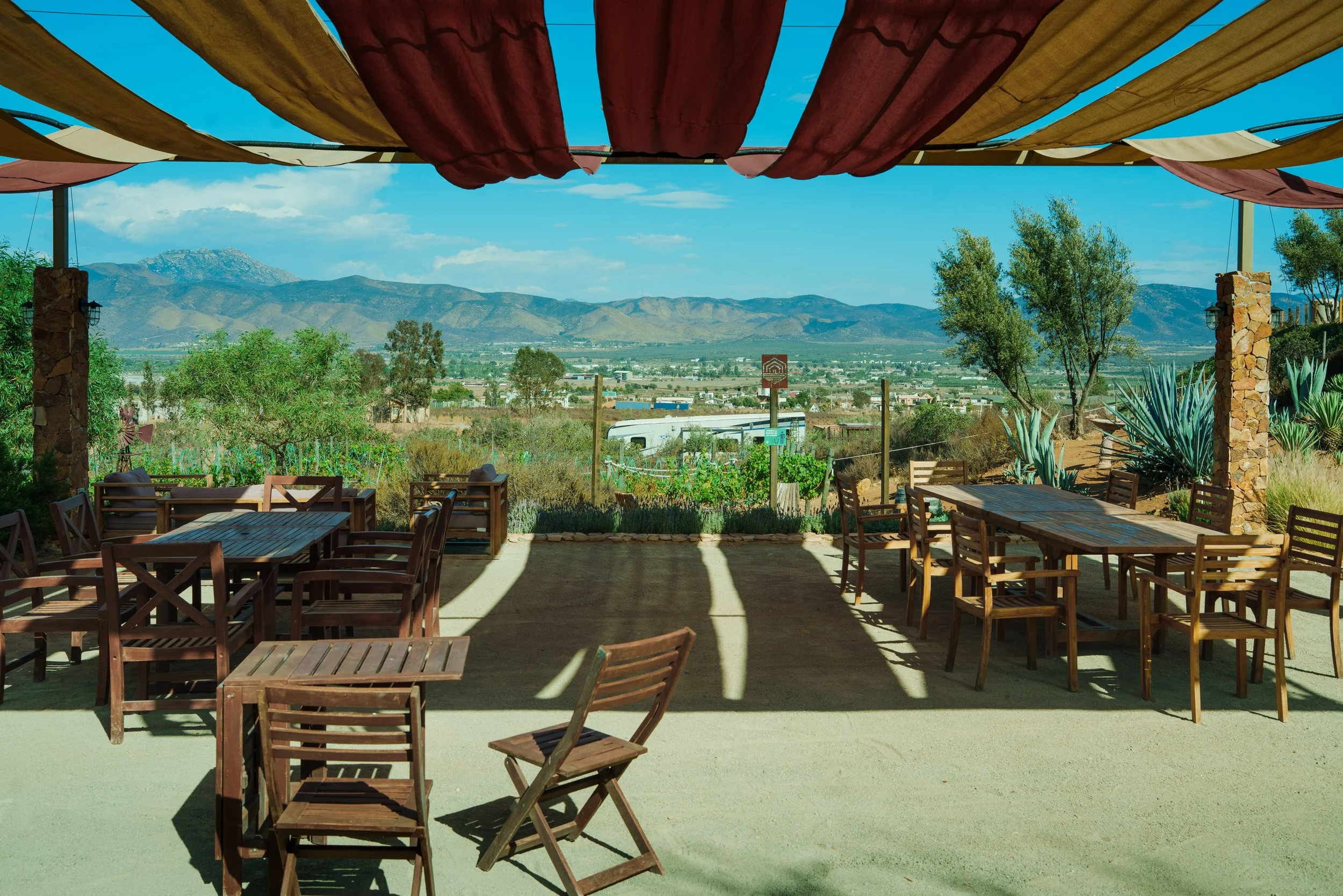 Outdoor patio with wooden tables and chairs, covered by a fabric shade, overlooking a valley with mountains in the background.