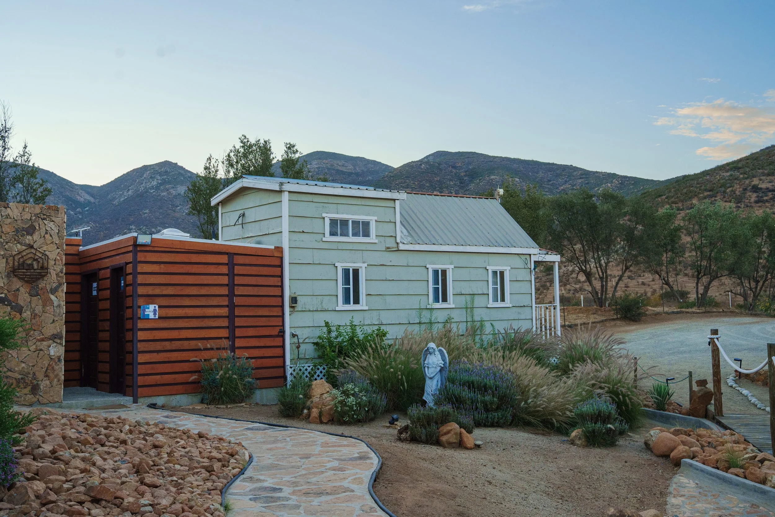 A house with light green siding and a metal roof is surrounded by a landscaped yard with a stone pathway, rocks, and various plants. There are hills or mountains in the background under a clear sky.