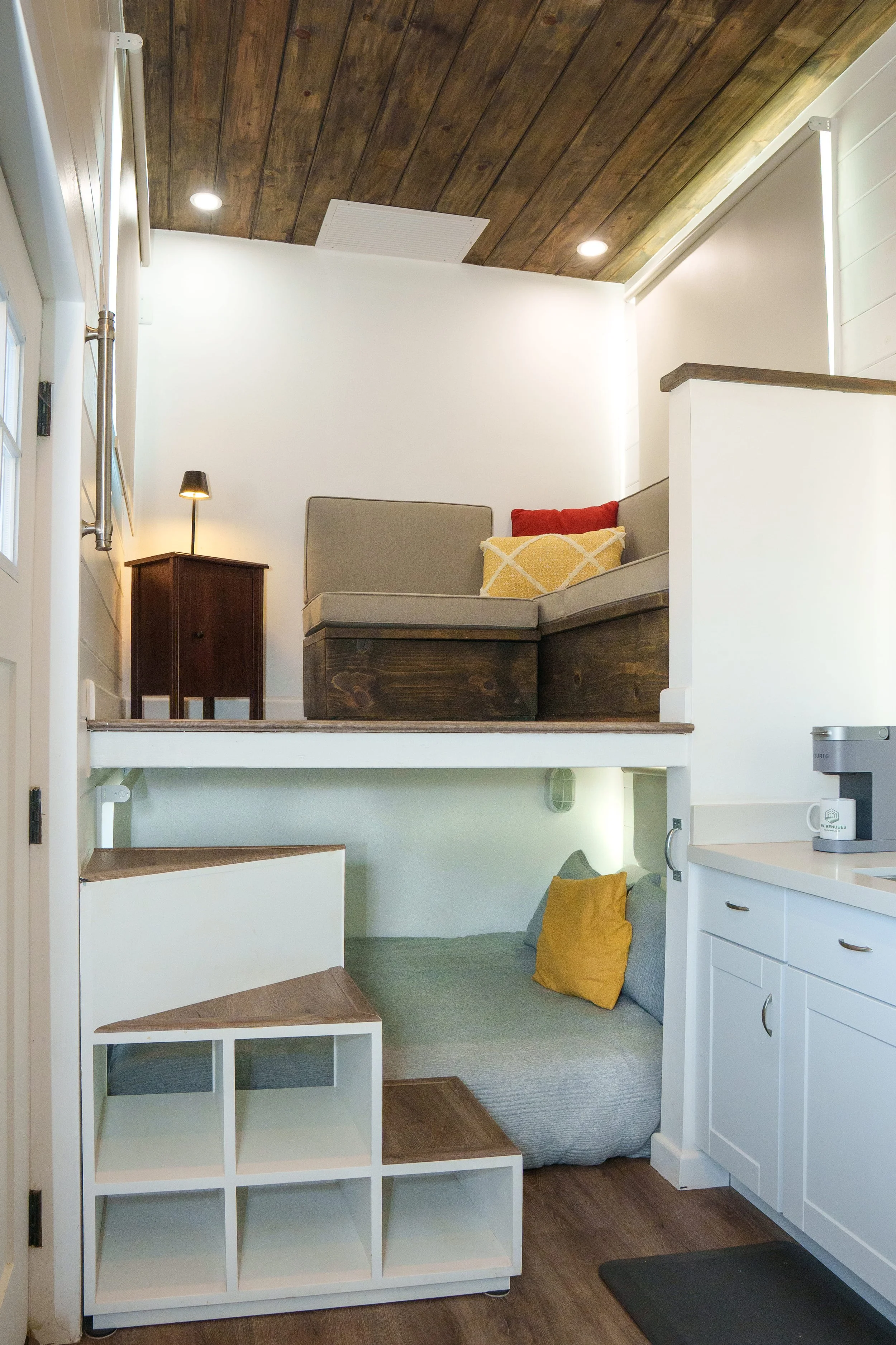 Interior view of a small living space with a wooden ceiling, white walls, and a cozy corner with pillows on the floor. There is a loft area with a beige couch and colorful throw pillows, a small wooden side table and a lamp. The lower area has a whit