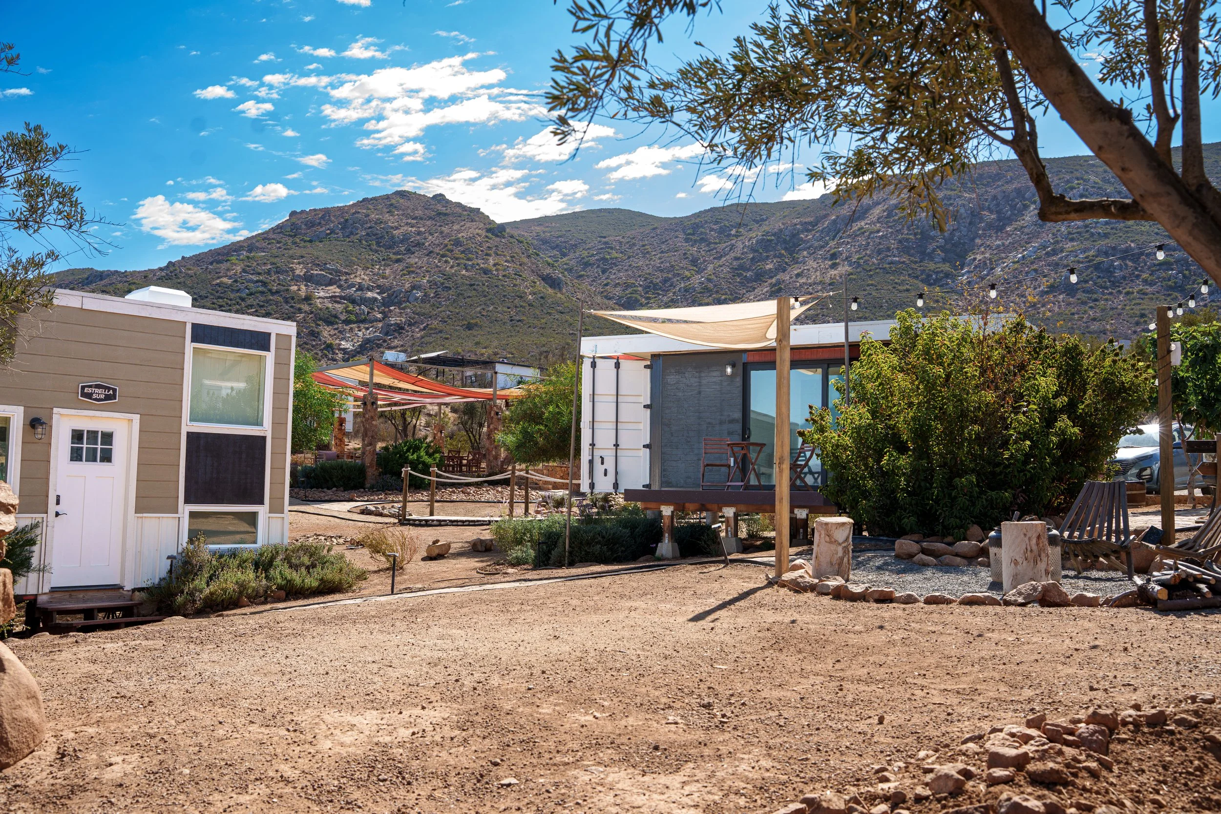 Outdoor scene with small modern buildings, string lights, trees, rocks, and a mountain in the background under a partly cloudy sky.