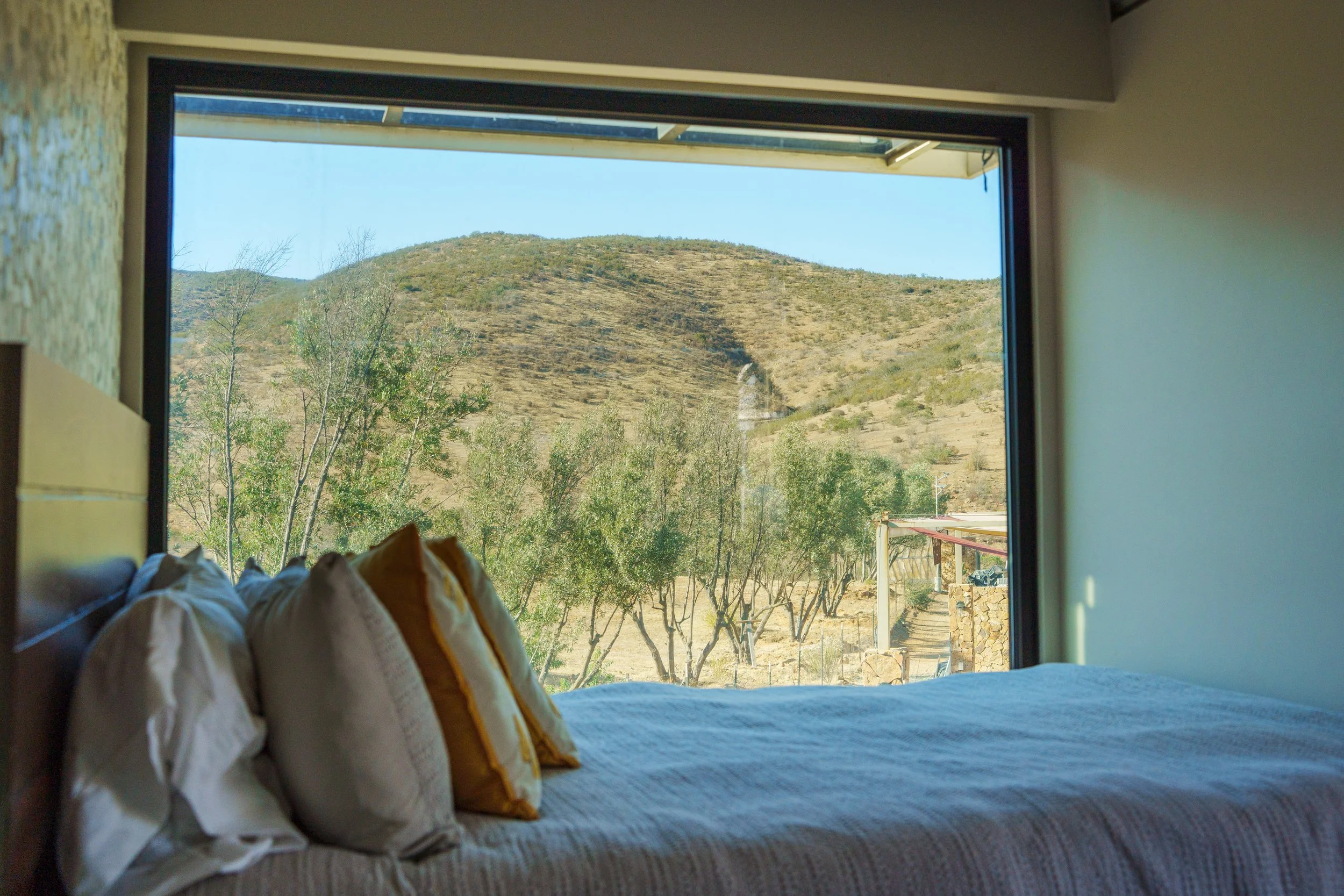 A bedroom with a large window showing a view of a arid hillside with sparse vegetation and a clear blue sky.