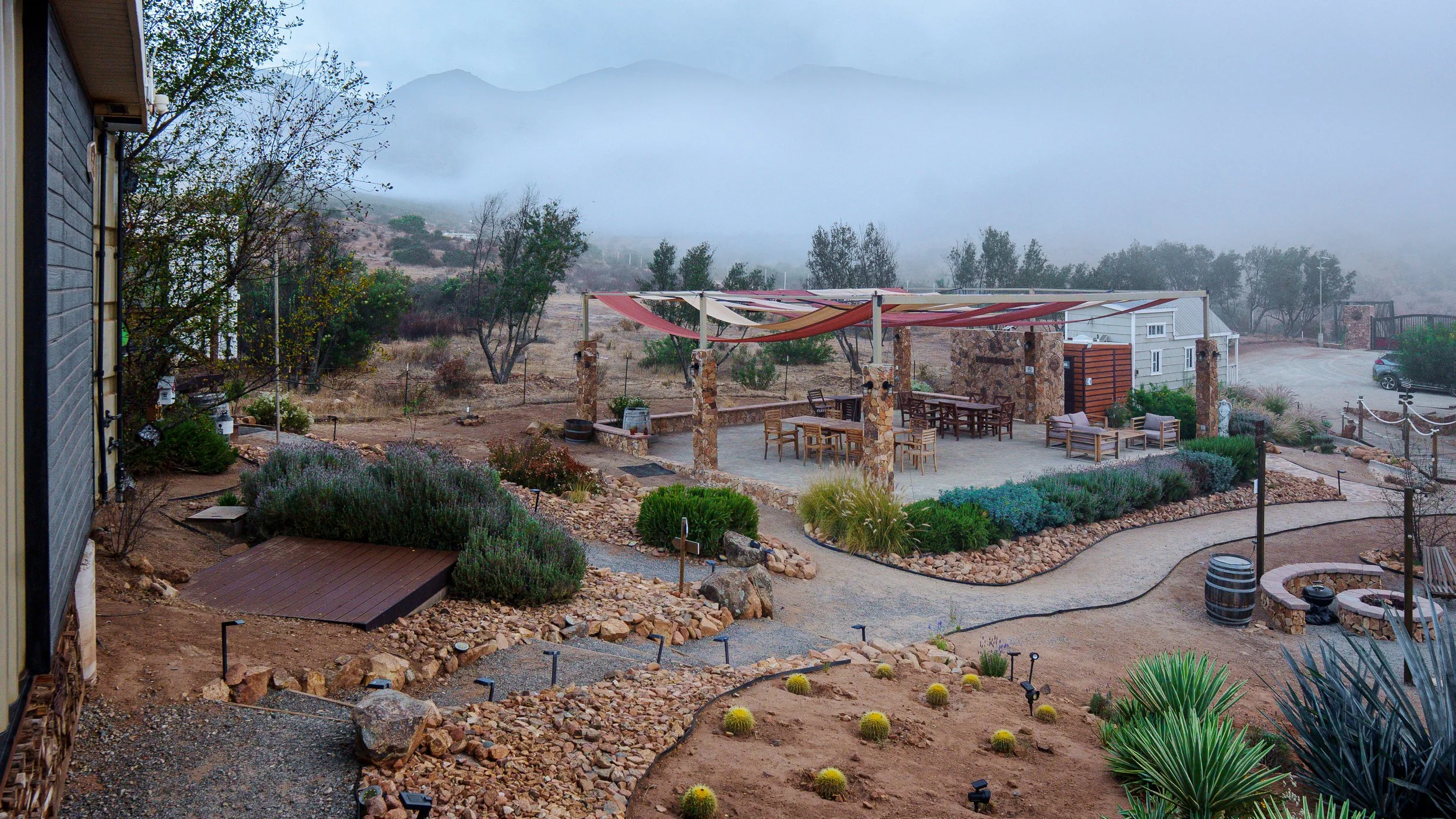 A misty outdoor patio area with tables and chairs, surrounded by desert plants, with a mountainous landscape in the background.