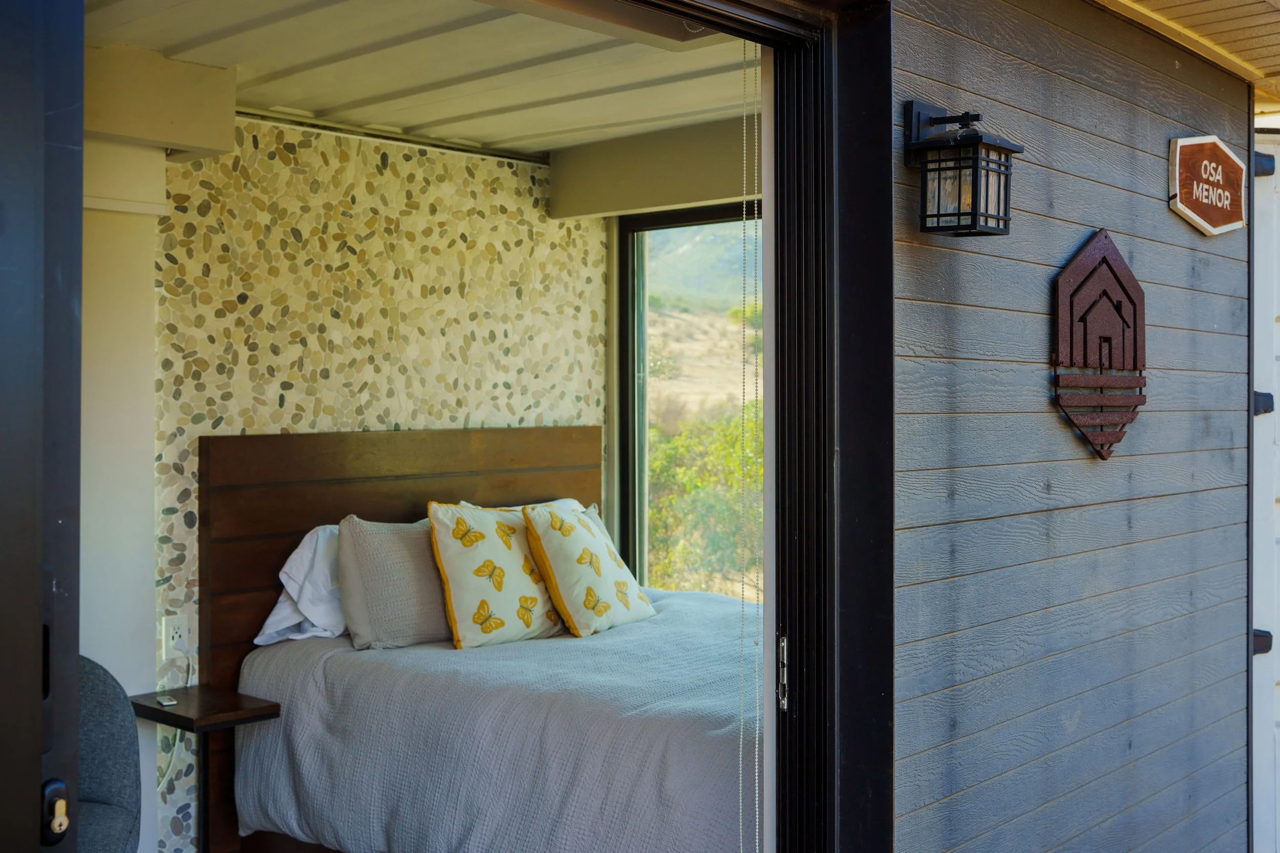 Interior of a bedroom with a window view, a bed with white bedding and yellow butterfly-patterned pillows, a wooden headboard, a side table, and a wall with a terrazzo pattern. Part of an exterior wall with dark horizontal wooden siding and decorativ
