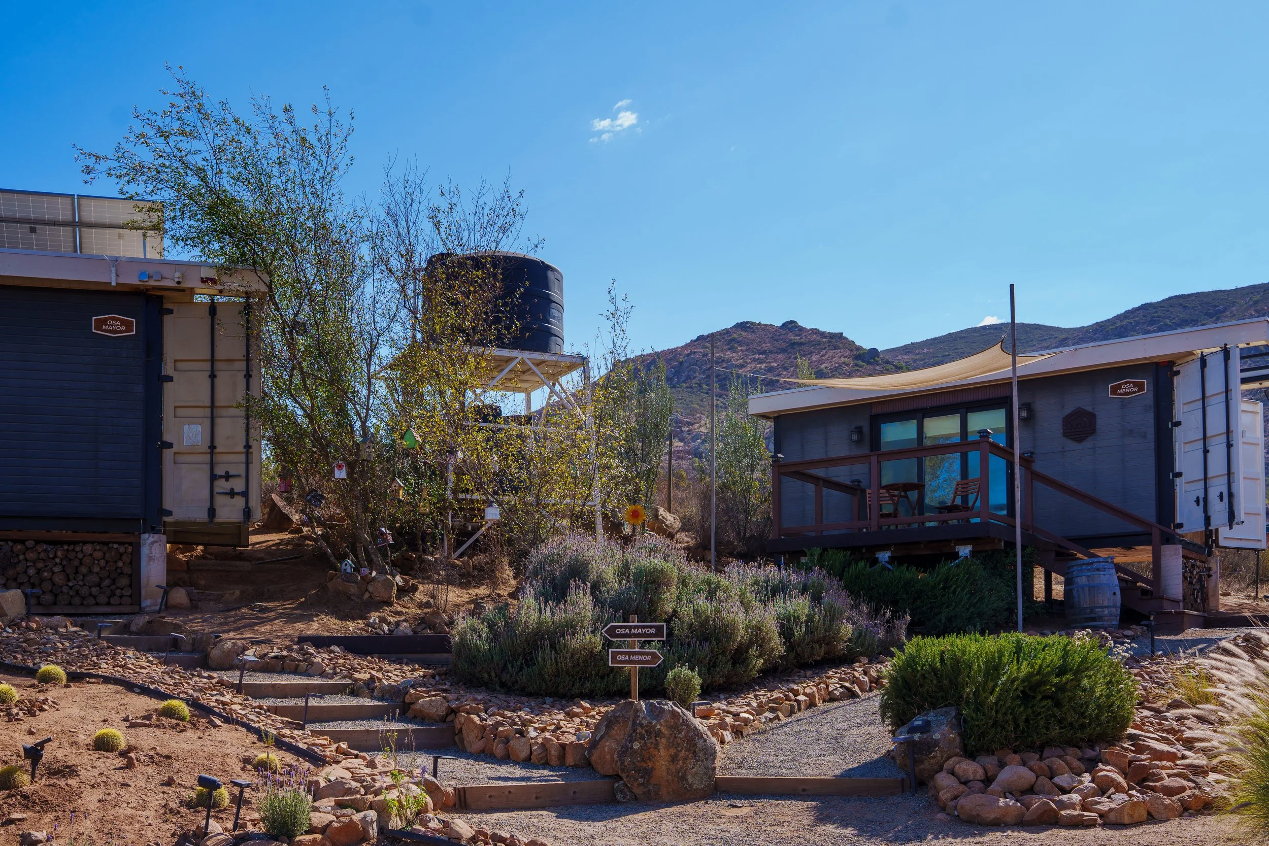 A landscaped outdoor scene with small buildings, trees, rocks, and garden signs, set against mountainous terrain and a clear blue sky.