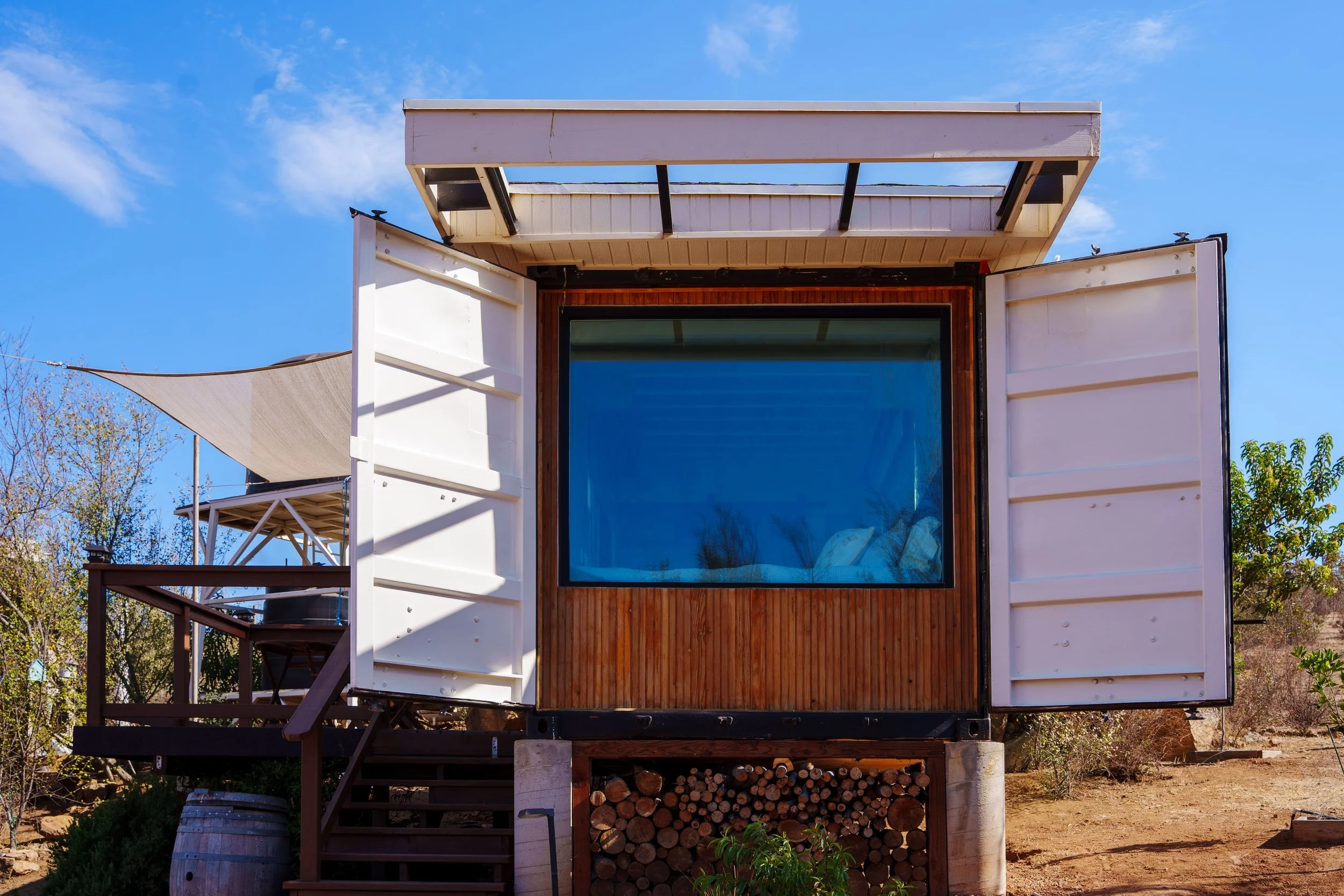 Modern tiny house with large window, white exterior walls, wooden accent wall, and storage for firewood below. It features an overhang and is set against a blue sky with some trees in the background.