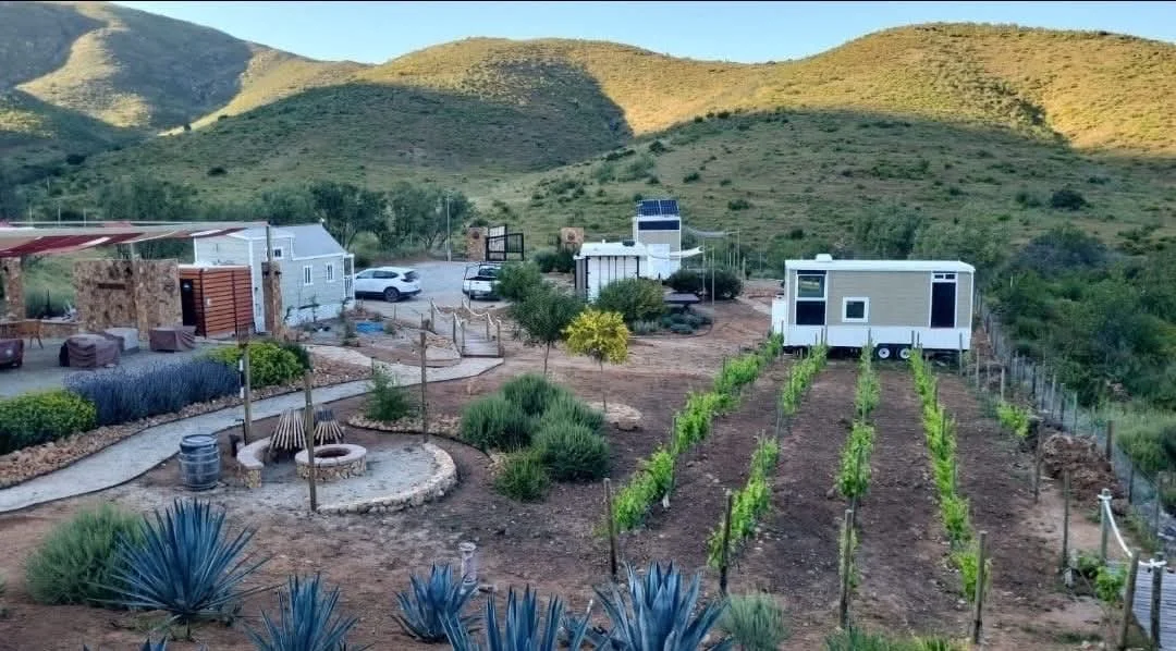 A rural landscape with hills in the background, featuring a small farm or homestead with garden beds, a seated area with a fire pit, a covered area, parked cars, and a tiny house or mobile home on wheels.