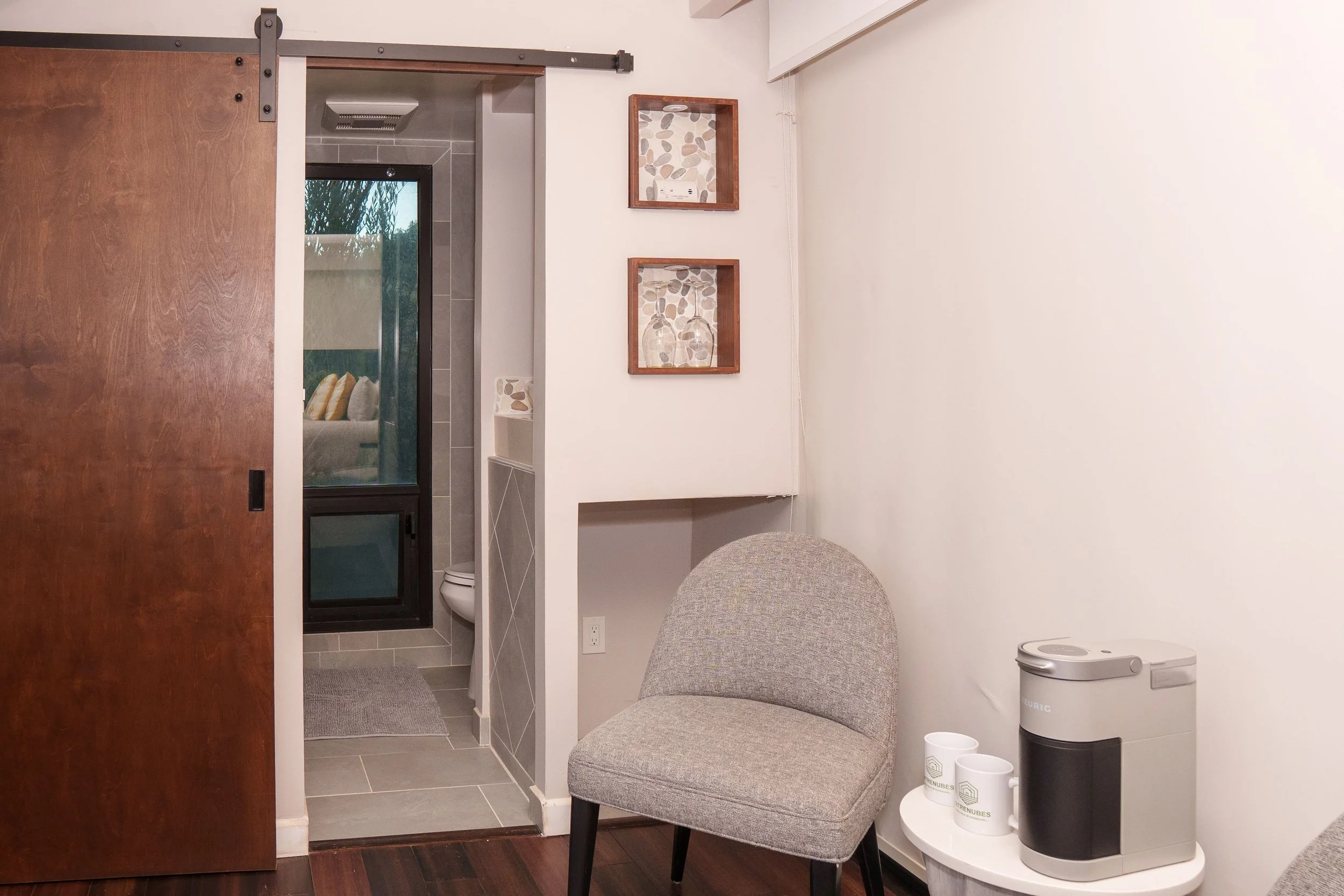 Interior view of a room with a wooden sliding door partially open, revealing a bathroom with a toilet, tiled floor, and a window. Outside the room, there are two upholstered chairs, a small table with a coffee machine, and wall decorations with pebbl
