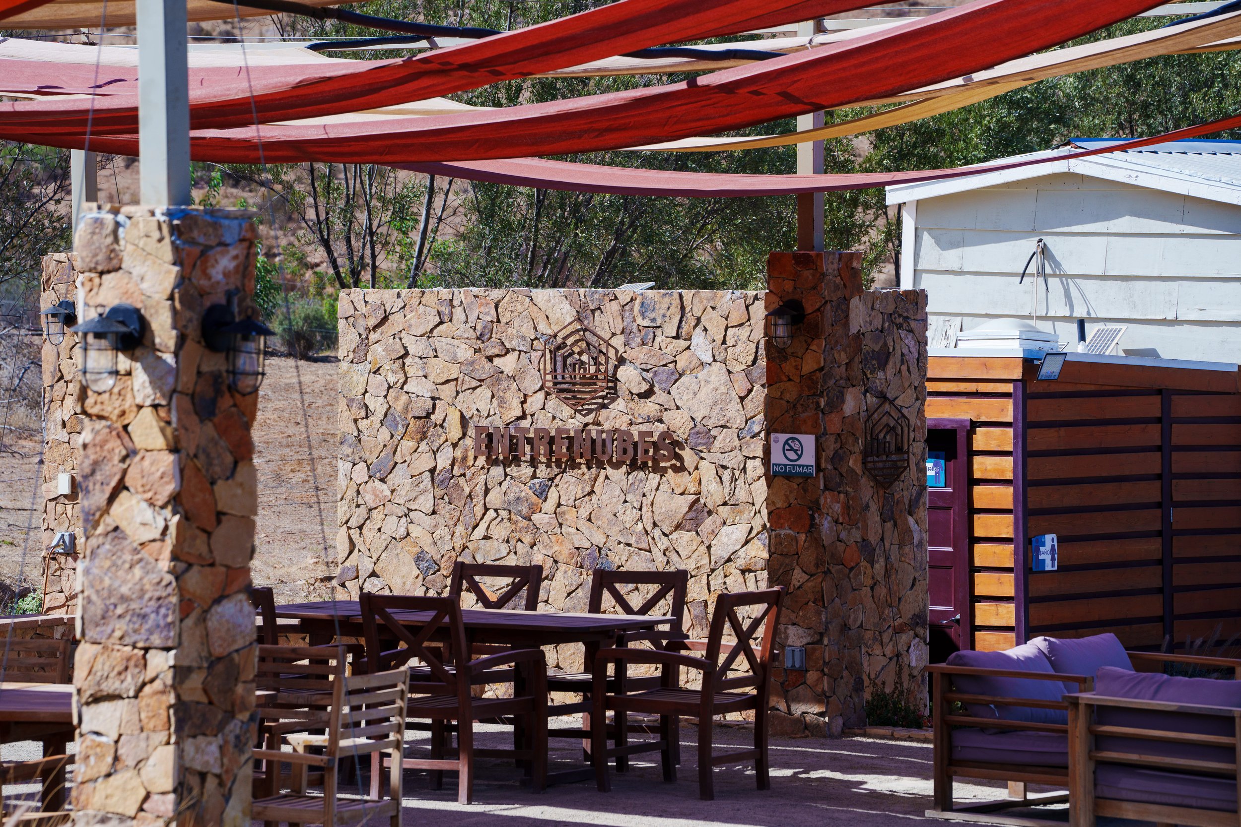 Outdoor seating area with multiple wooden tables and chairs, a stone entrance with a sign that says "ENTRENUBES," and a white building in the background. Shade cloths overhead and trees around the area.