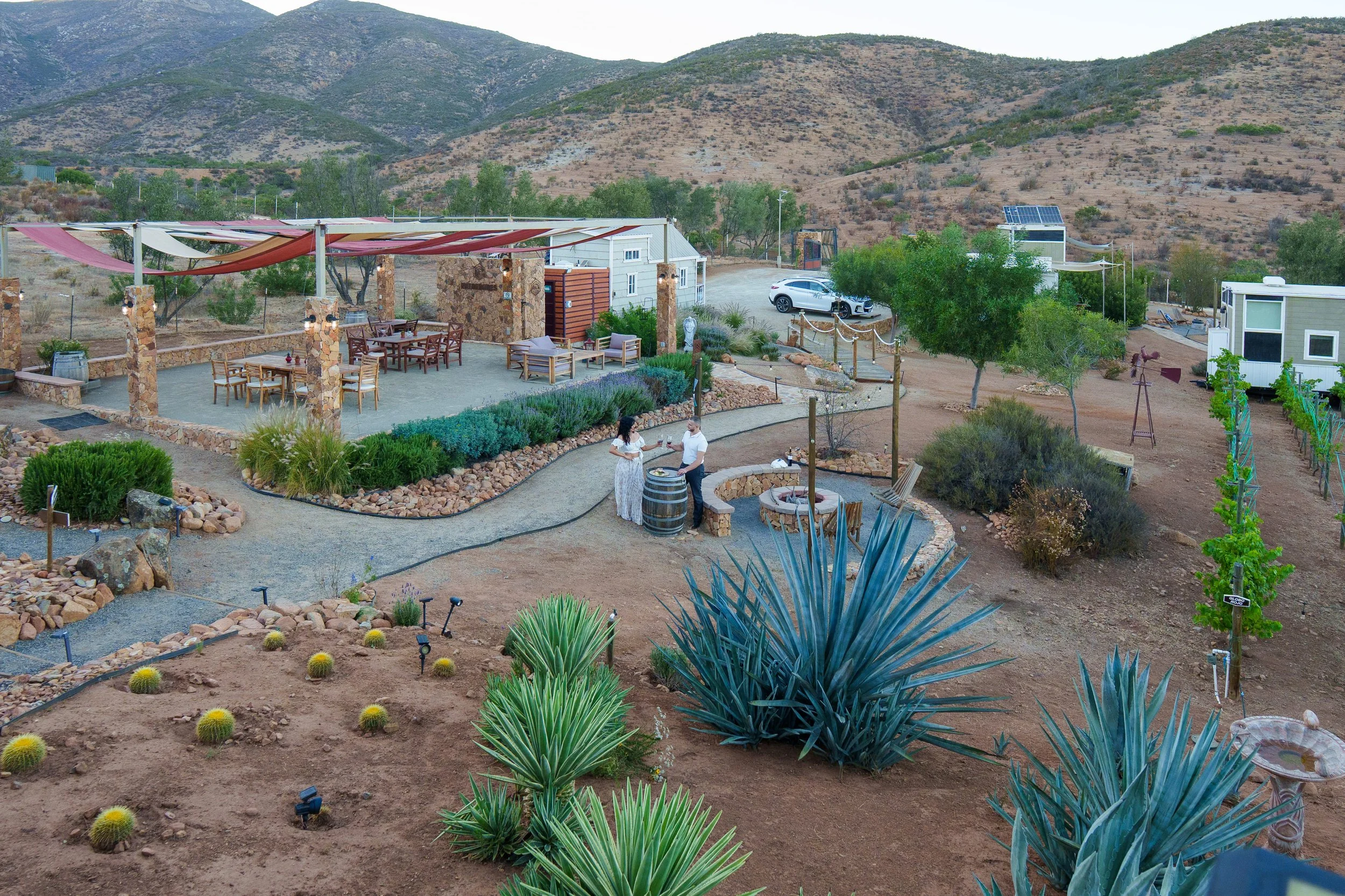 A desert landscape features a patio with outdoor seating, two people socializing near a barrel table, various plants including agave and cacti, a gravel path, and a scenic mountain backdrop with sparse vegetation.