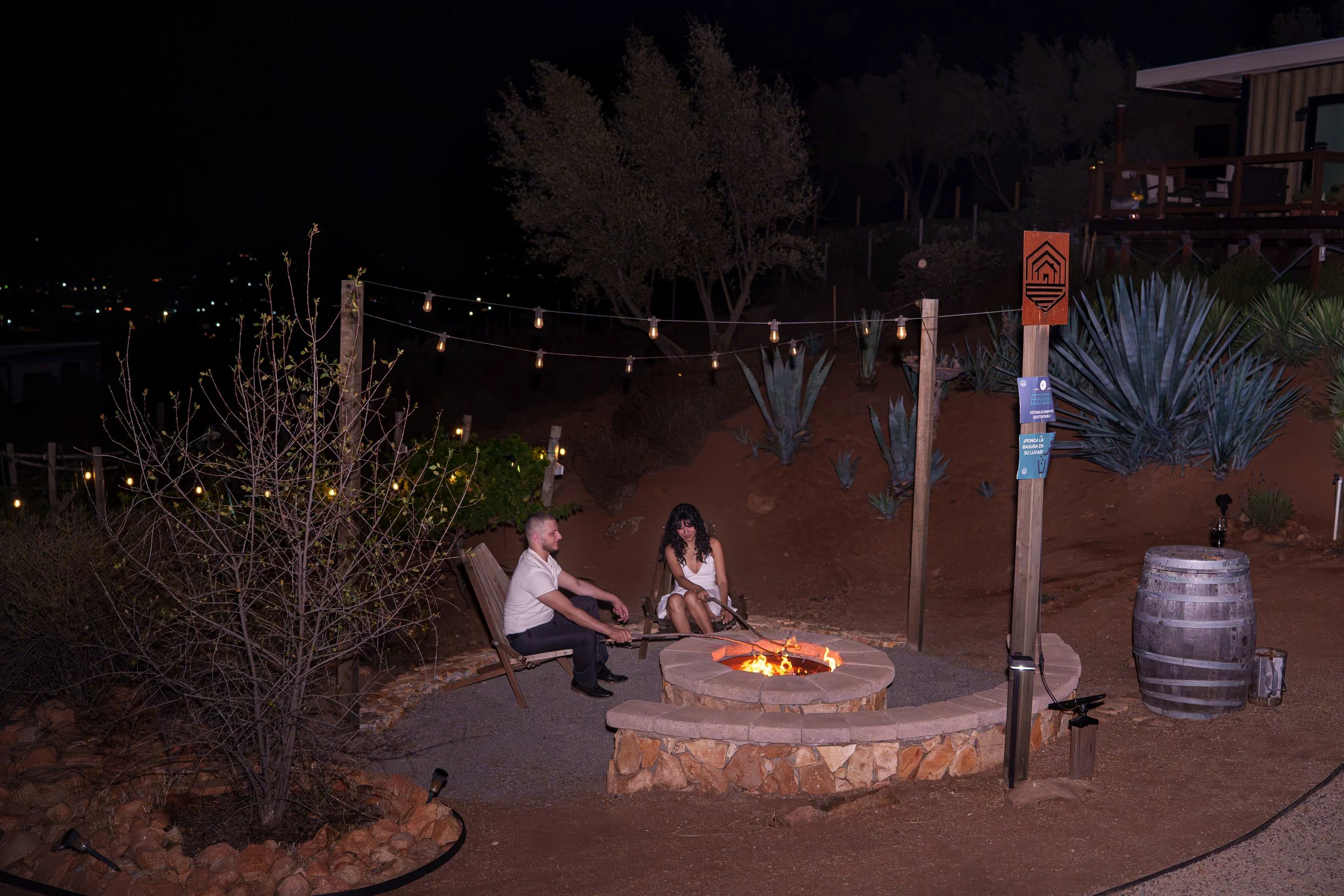 Nighttime outdoor gathering around a fire pit with two people sitting in wooden chairs, string lights overhead, desert plants, a wooden deck with furniture, and a small sign on a post.