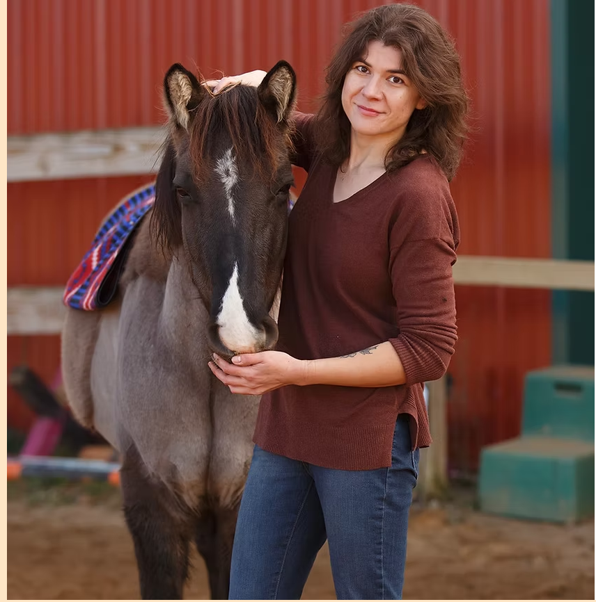 A woman, a telehealth therapist, in a brown sweater standing next to a brown and white horse, holding its nose. The woman has curly hair and is smiling. The background features a red wooden fence and some structures.