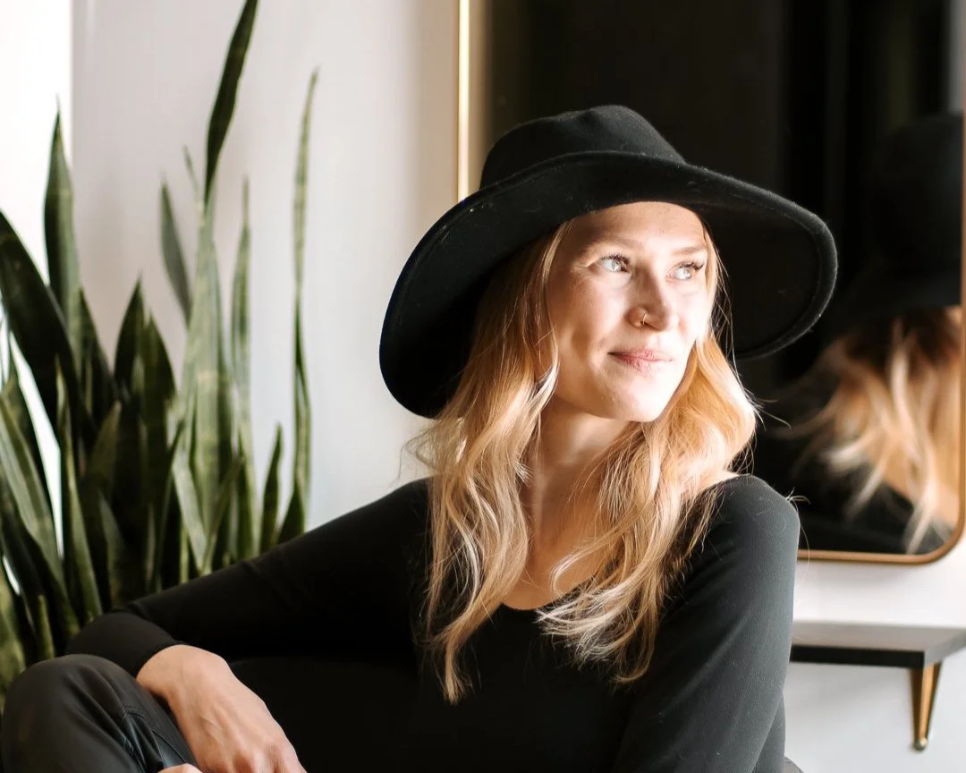 A woman with wavy blonde hair wearing a black wide-brimmed hat and black top sitting near a mirror, with a plant in the background.