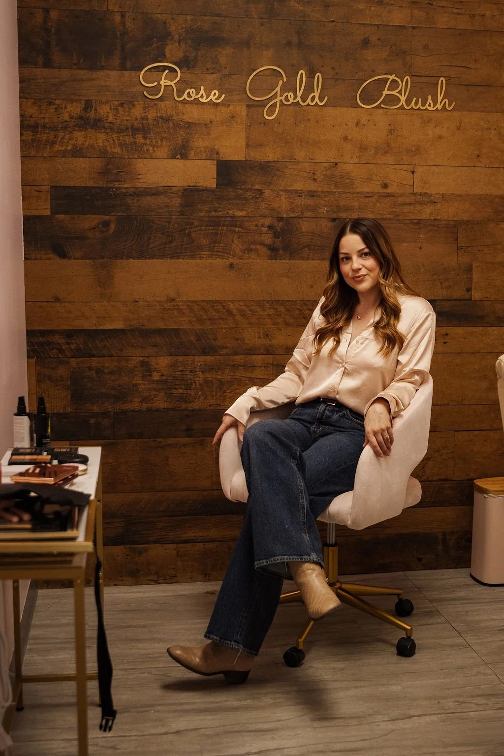 A woman with long, wavy blonde hair, wearing a bright pink blazer and white top, smiling and holding a makeup brush.