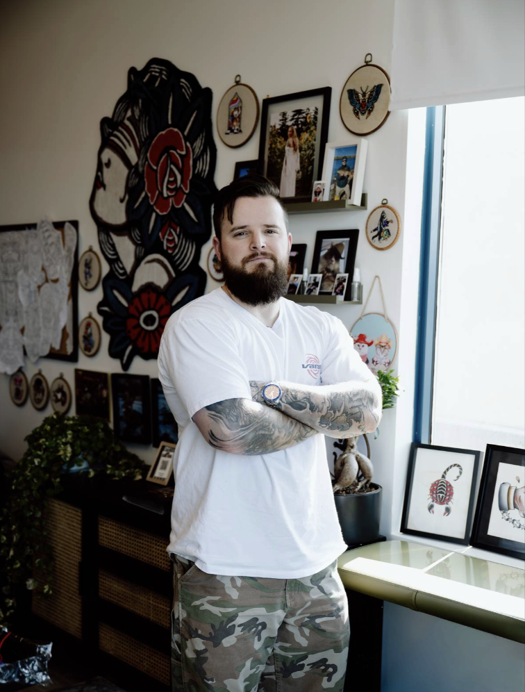 A smiling man with a beard and tattoos on both arms standing in front of a brick wall, wearing a black T-shirt featuring an image of a basketball player with the number 23 and the name 'Jordan'.