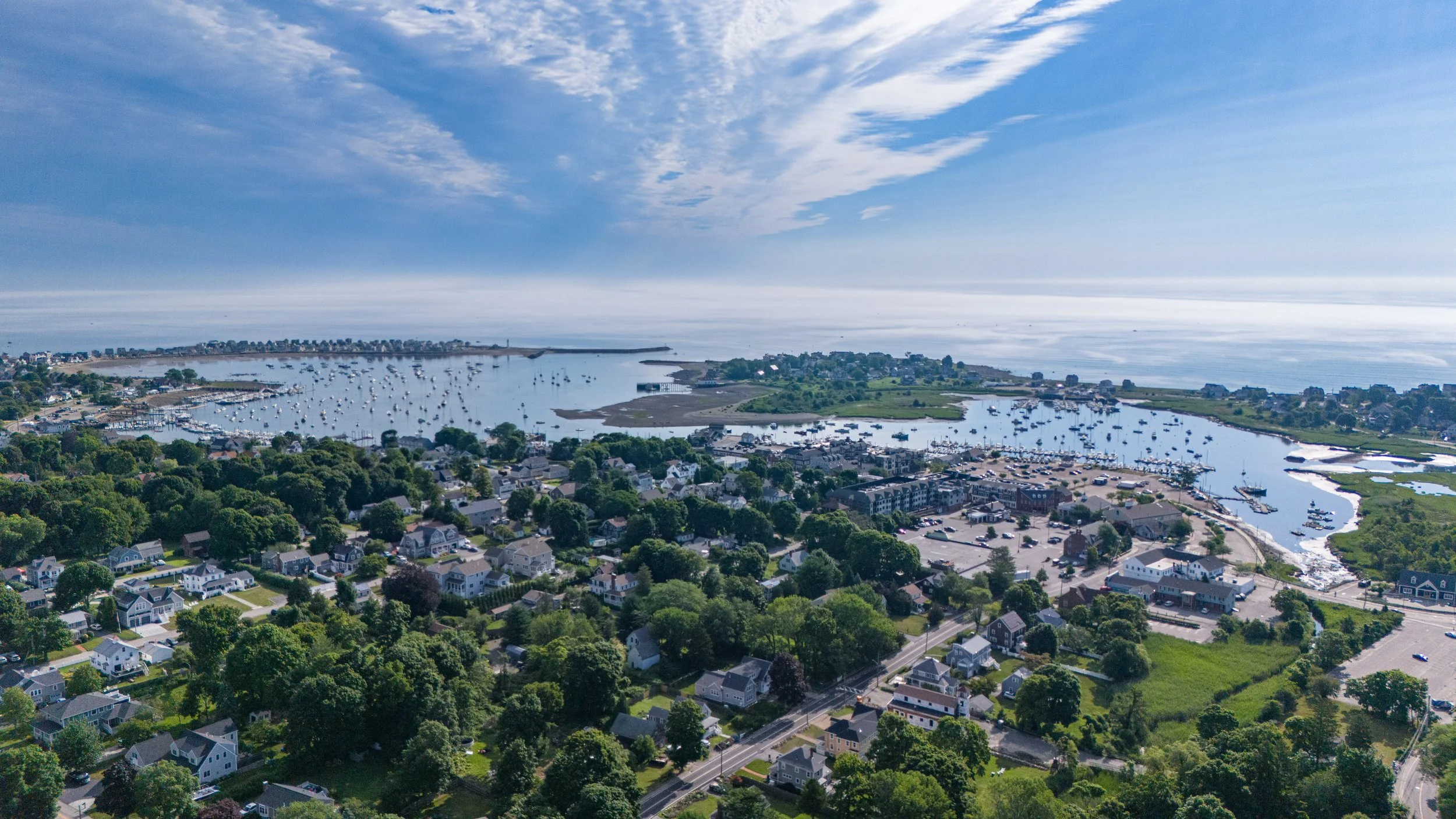 Aerial view of a coastal town with houses, a marina filled with boats, green trees, and a body of water under a partly cloudy sky.