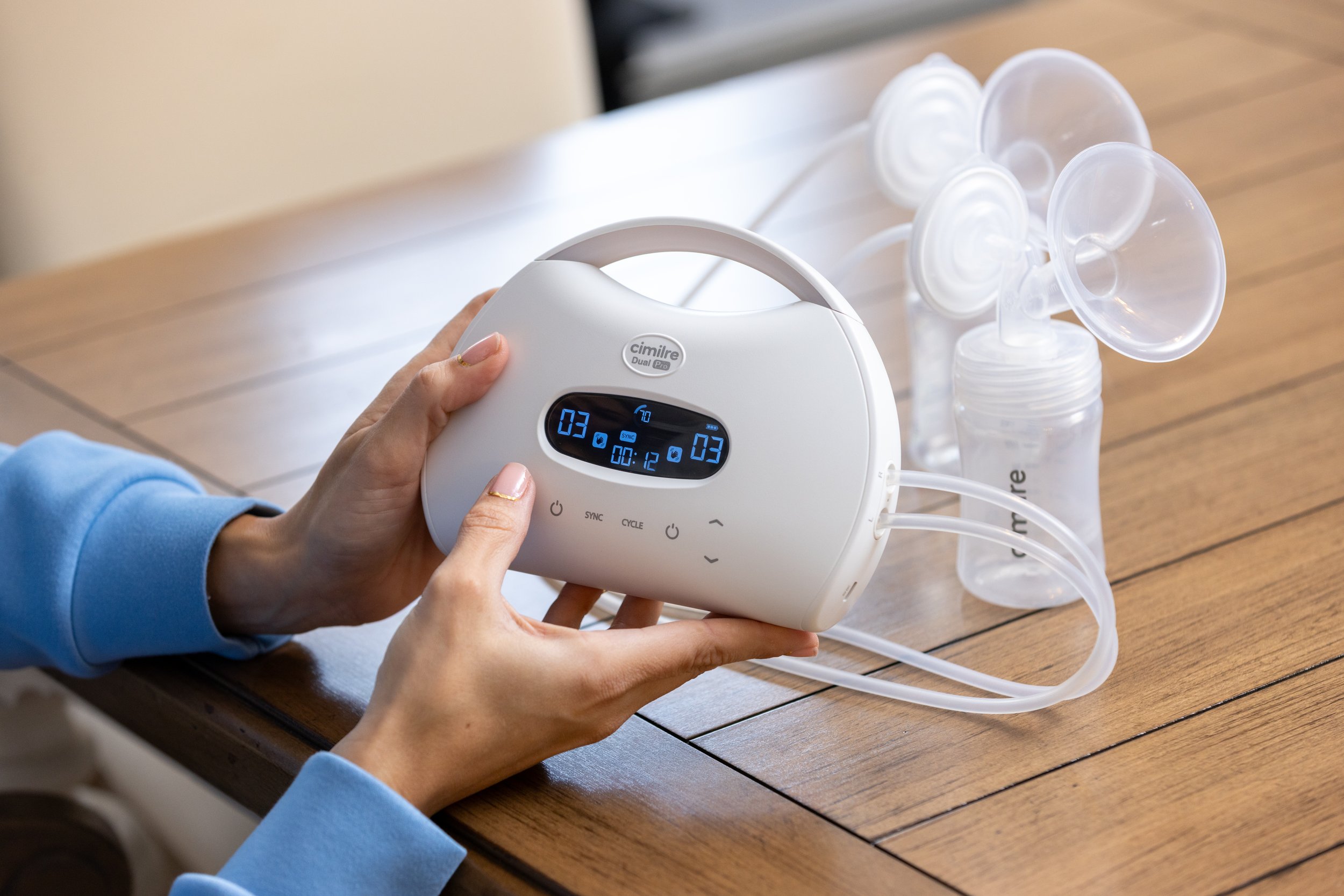 Person operating a Cimilre DualPro breast pump on a wooden table with milk collection bottles attached.