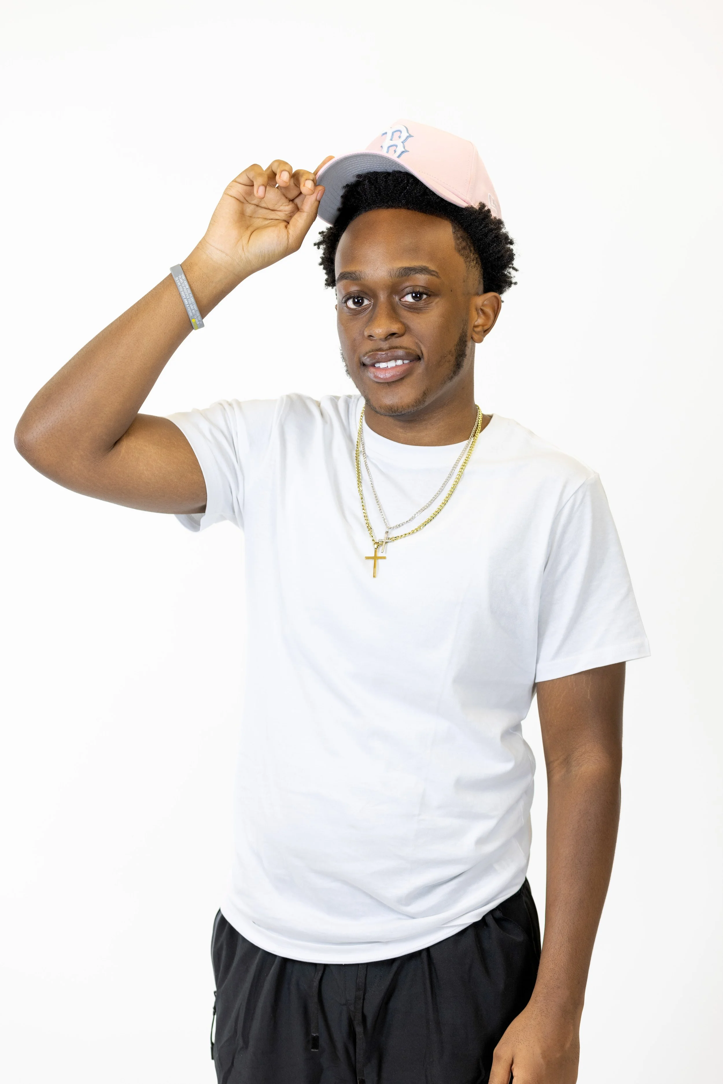 Young man wearing a white t-shirt, gold chains, and a pink baseball cap, holding the cap brim with one hand, smiling against a plain white background.