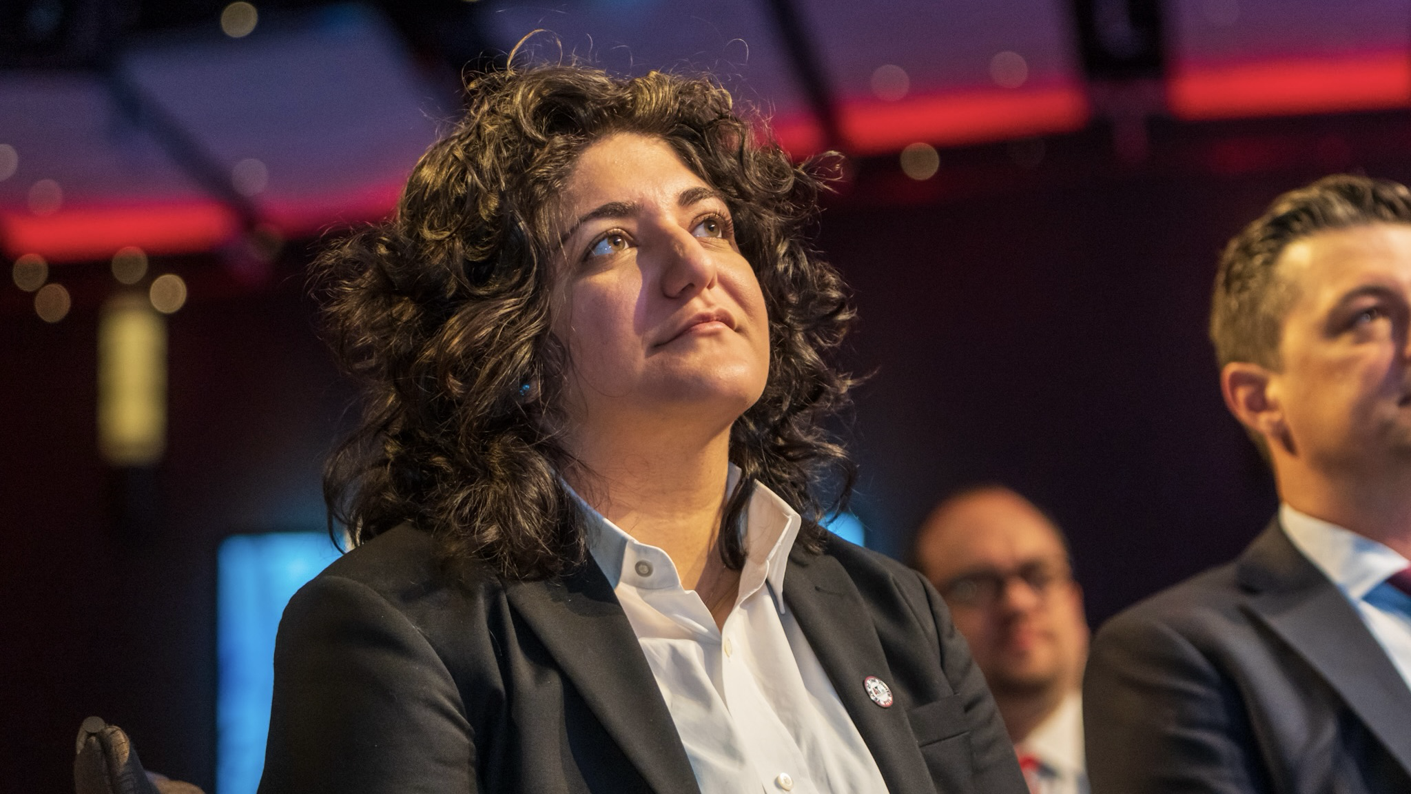 council member Liliana Bakhtiari in a black blazer and white shirt seated in an auditorium, looking upward under colorful stage lighting