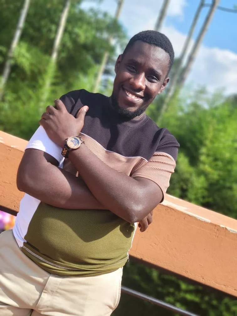 Smiling young man standing outdoors on a sunny day, crossing his arms, wearing a striped short sleeve shirt and beige pants, with a watch on his wrist, and a wooden railing behind him with green trees and a partly cloudy sky in the background.