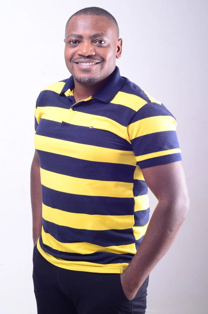 A smiling man with short hair and a beard wearing a yellow and navy blue striped polo shirt, standing with his hands in his pockets against a plain white background.