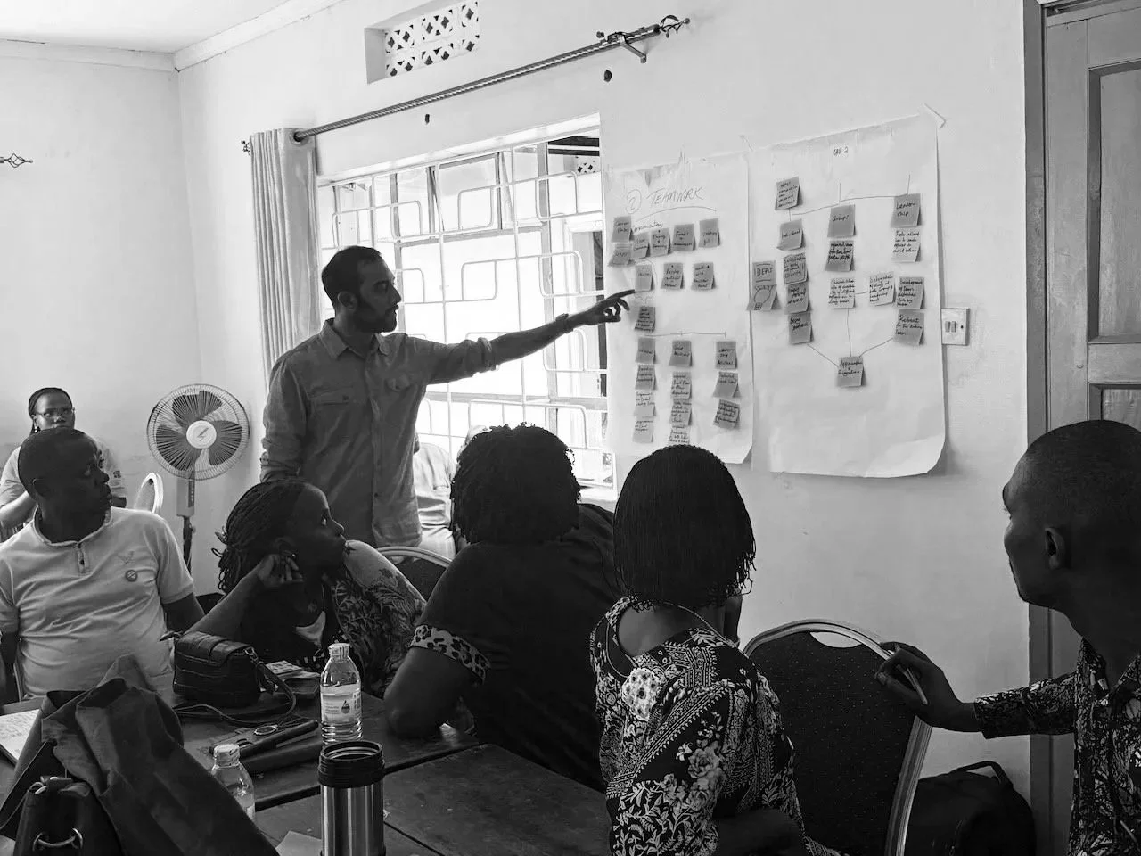 A man presents to a group of women seated around a table in a meeting room with white walls. The man points to a large paper with sticky notes on the wall, and the women listen attentively. The room has a window with curtains, a fan, and various items on the table.