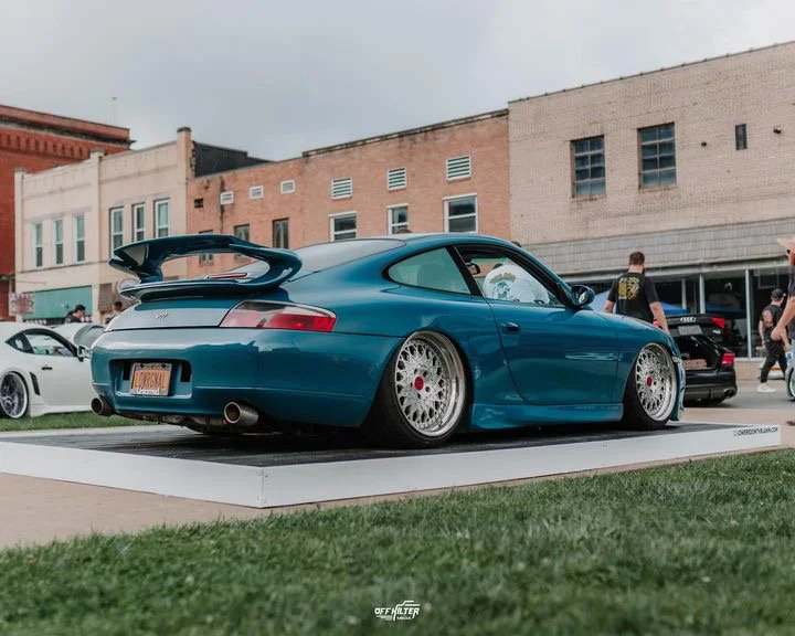 Blue sports car on display platform, with modified rims and spoiler, parked outdoors at a car show.