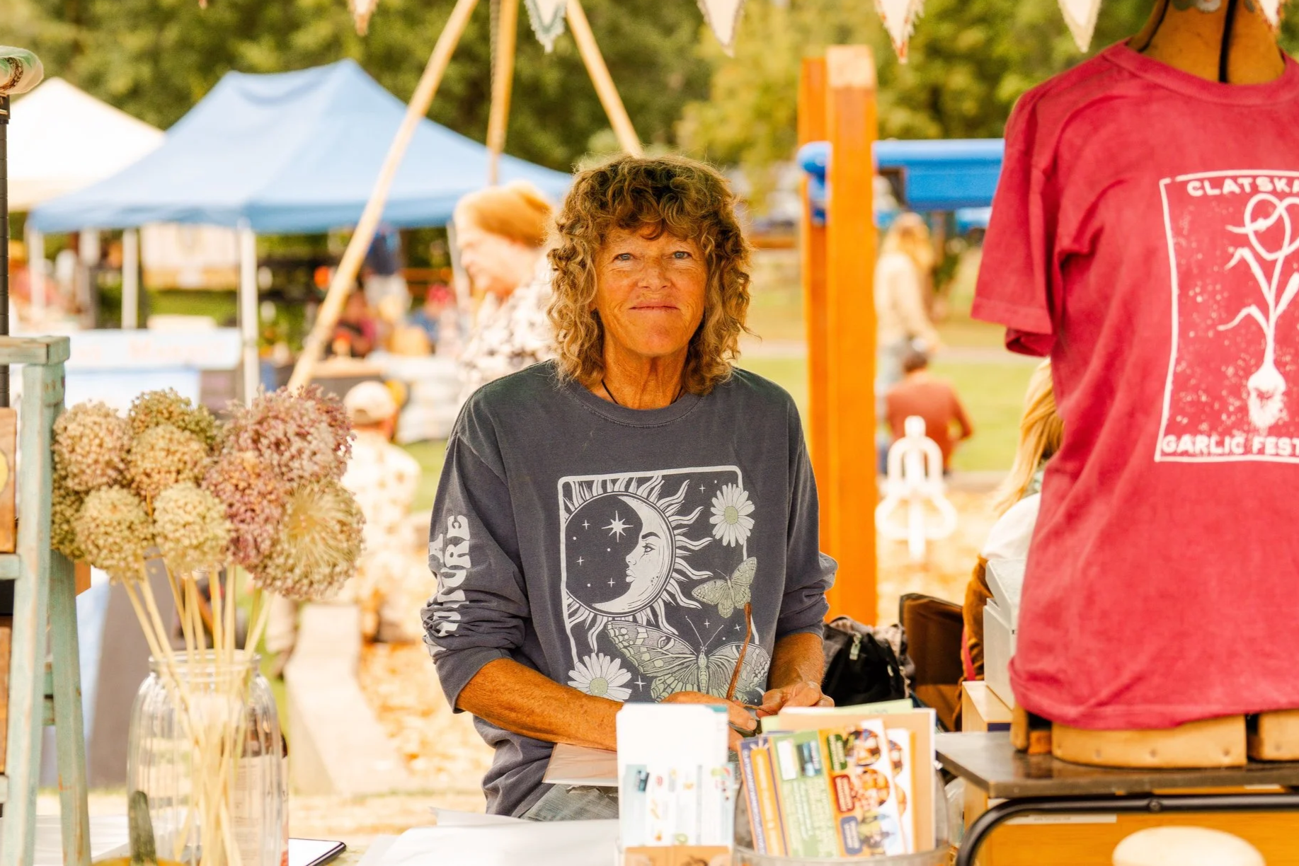 A smiling woman with curly hair wearing a jacket and glasses on top of her head, sitting indoors.