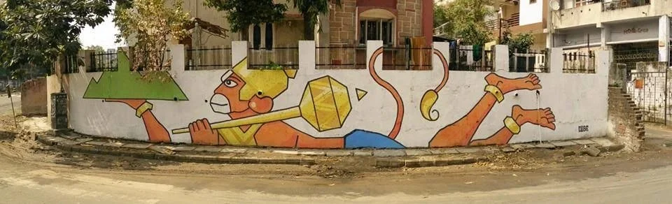 Colorful mural of a stylized god Hanuman carrying a Mountain, with exaggerated features, set against a white wall in an urban environment.