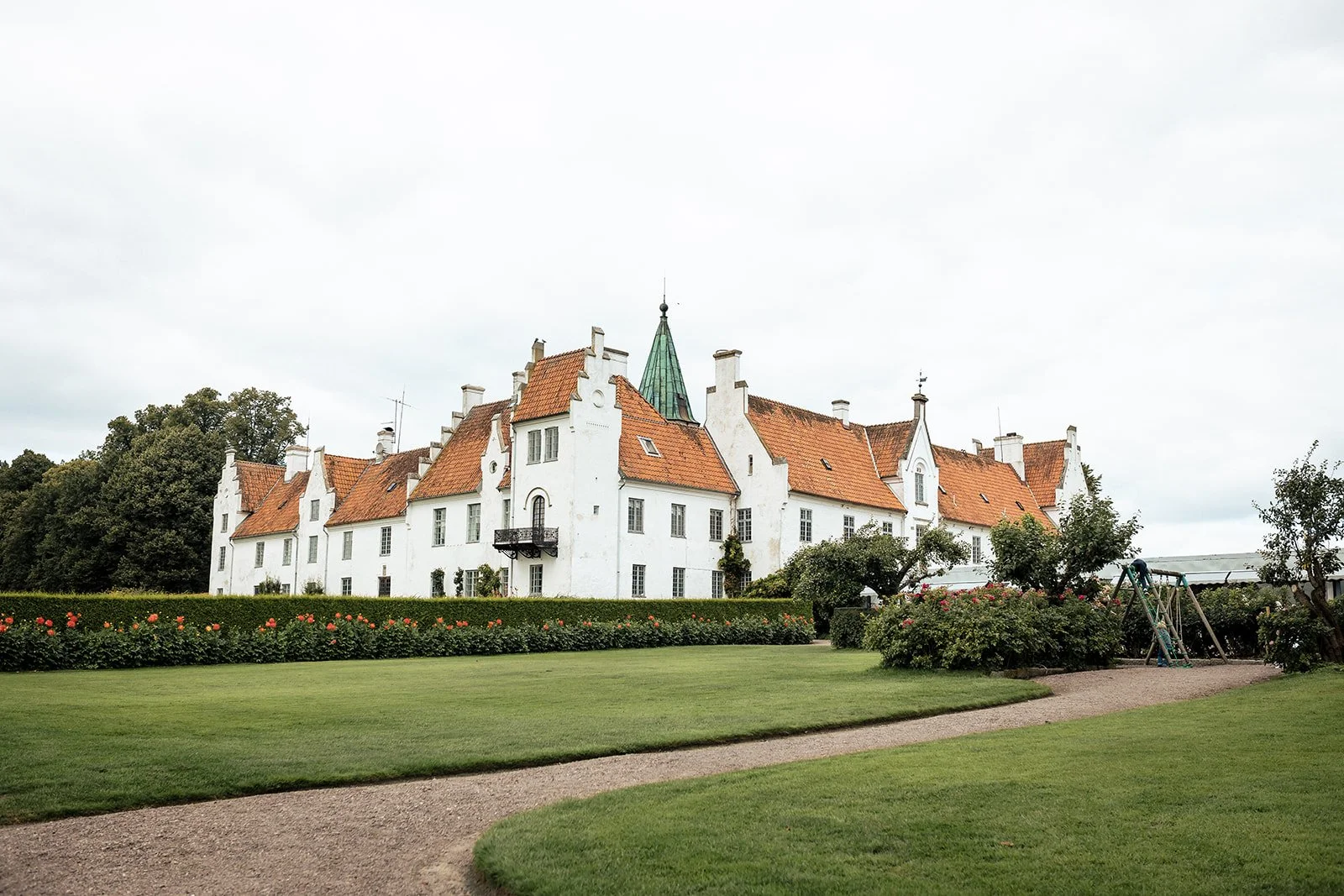 A large white historic house or castle with red-tiled roofs and a green spire, surrounded by a well-maintained garden with a walking path, bushes, and flowering plants.