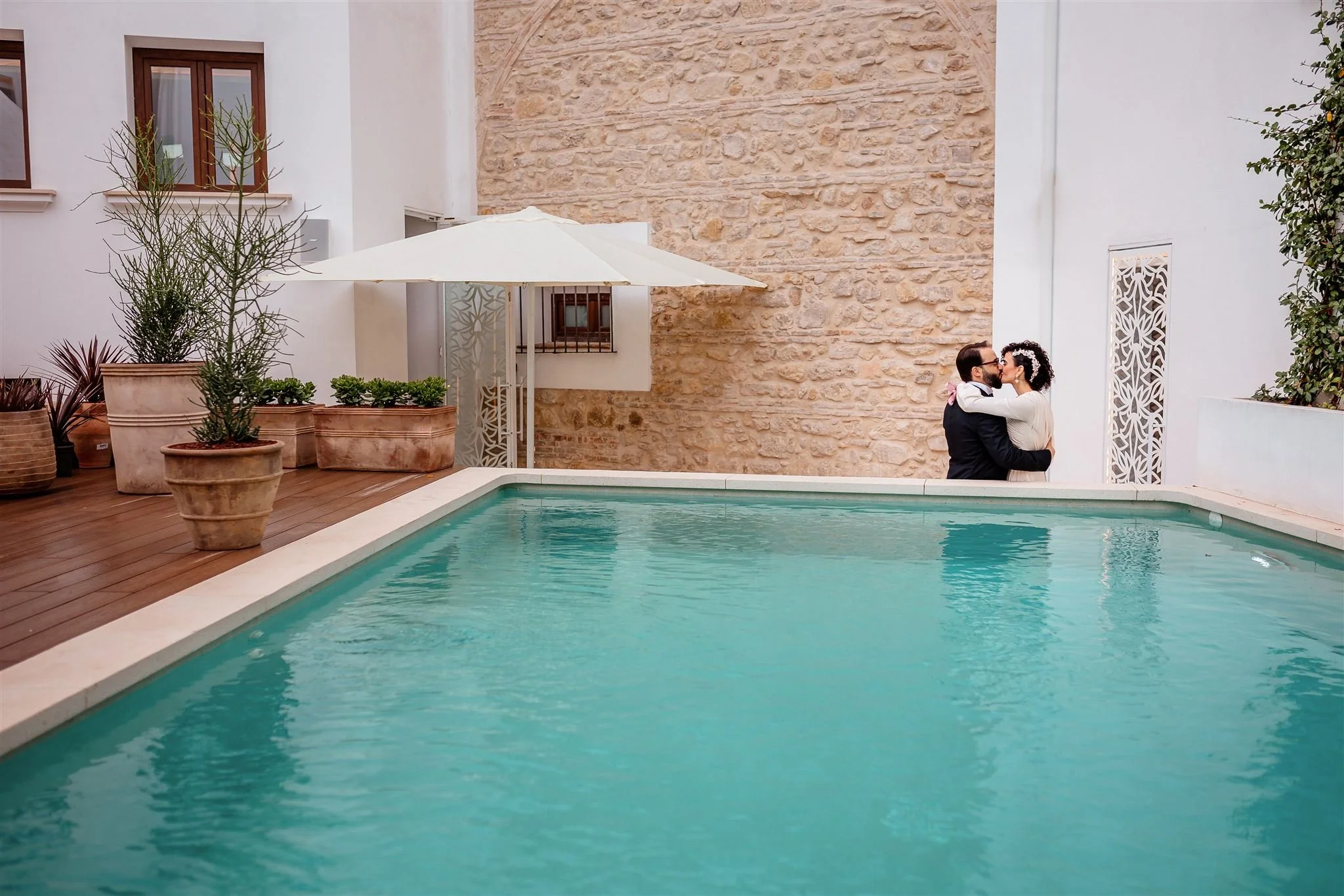 A couple embraces and kisses near a swimming pool in a courtyard with potted plants, a white umbrella, and a stone wall.