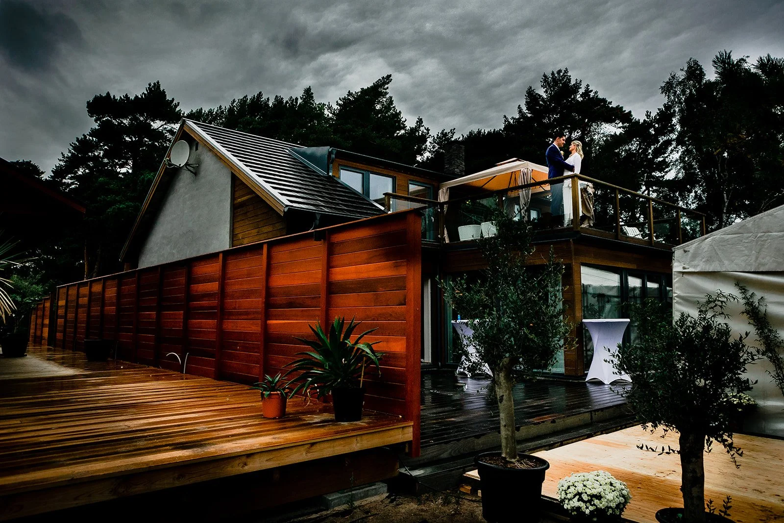Couple standing on a house's balcony during cloudy weather, with a wooden deck and potted plants in the foreground.