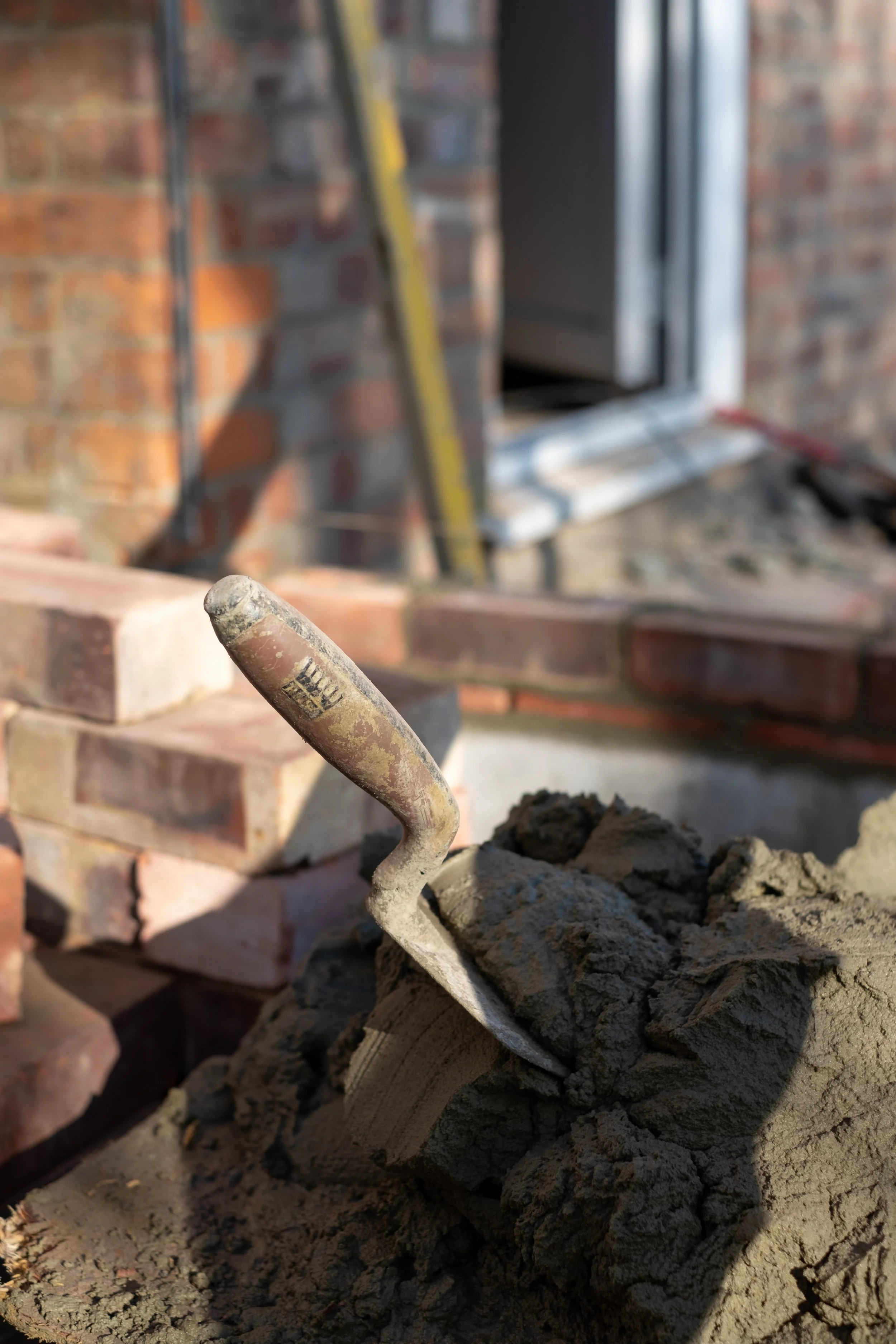 A trowel stuck in a pile of concrete or mortar with a brick wall and ladder in the background.