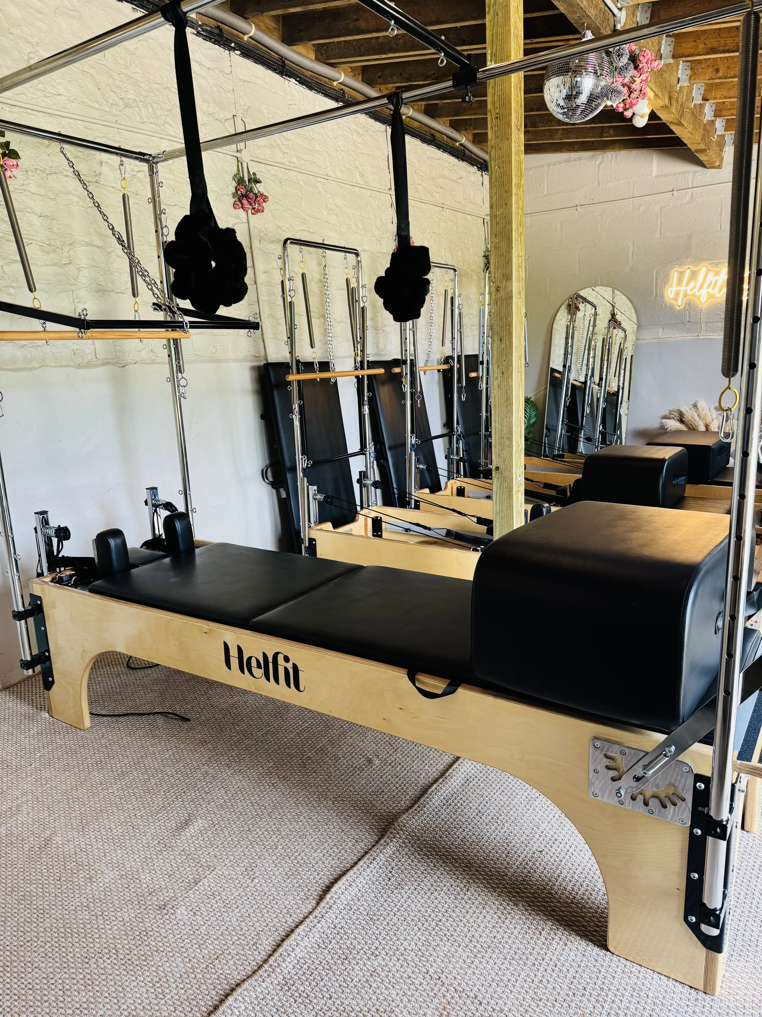 A Pilates reformer gym machine with a wooden frame, black padded platform, and various metal bars and straps, in a room with white brick walls, a mirror, hanging pink flowers, and a disco ball.