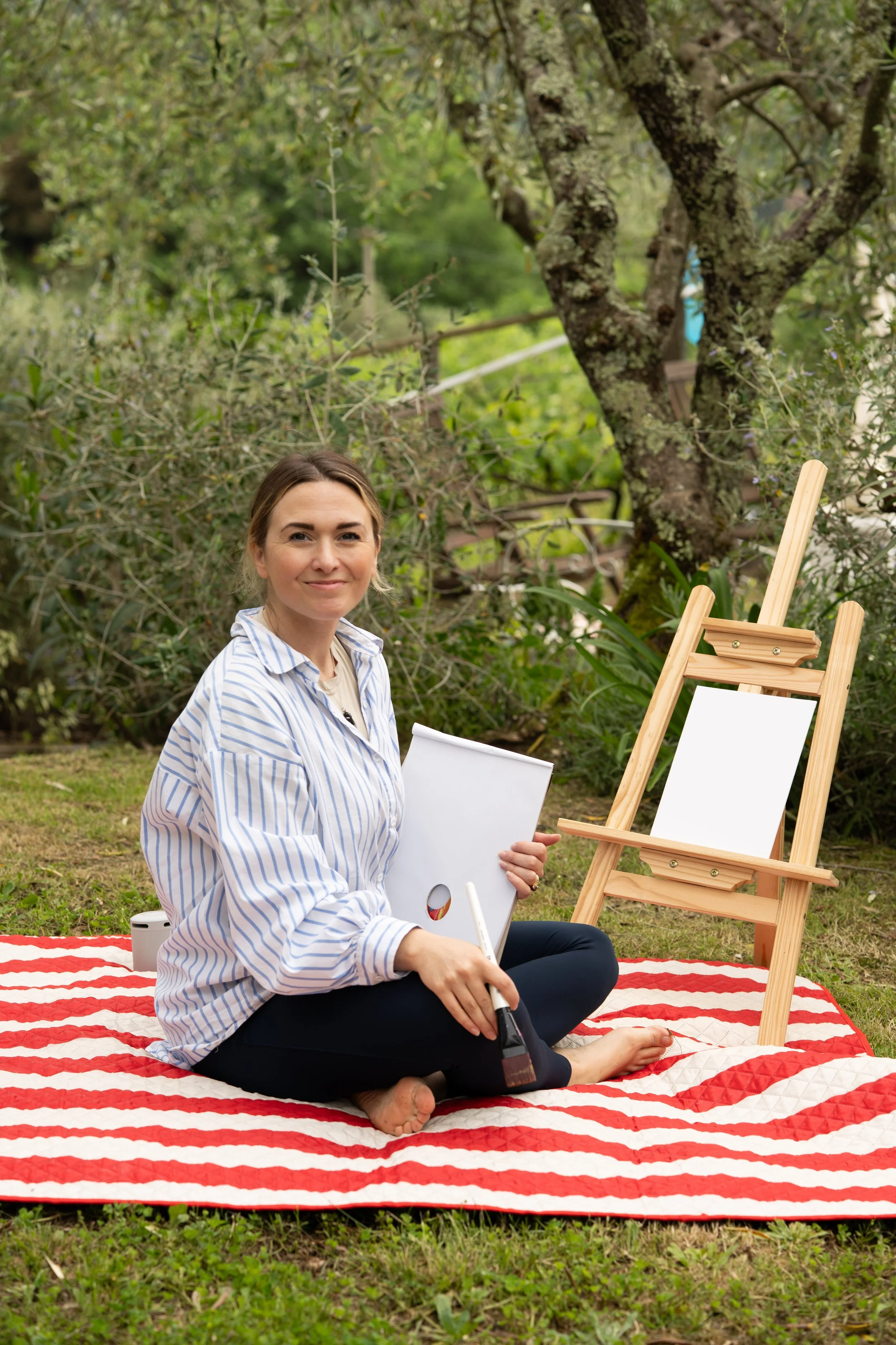 A woman sitting on a red and white striped blanket outdoors, holding a notebook and paintbrush, with an easel and greenery in the background.