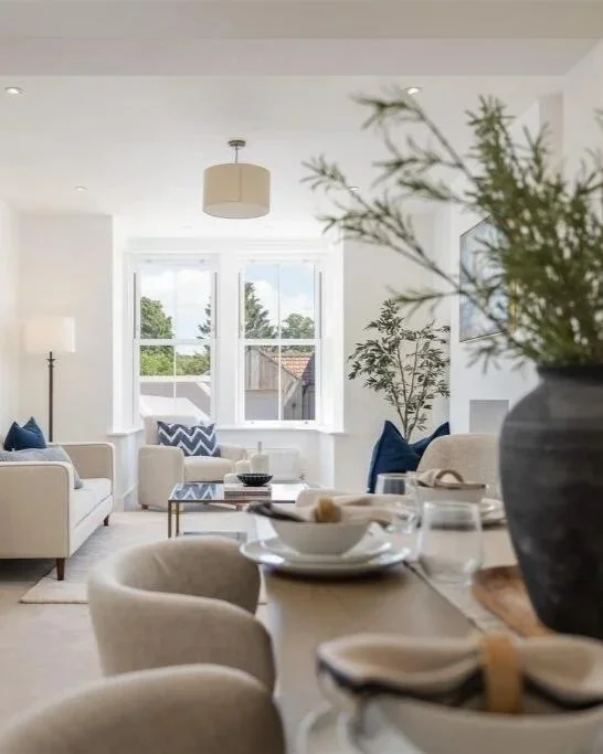 Bright living room with white walls, large windows, beige sofas with blue pillows, indoor plants, and a dining table set with white dishes and glassware.