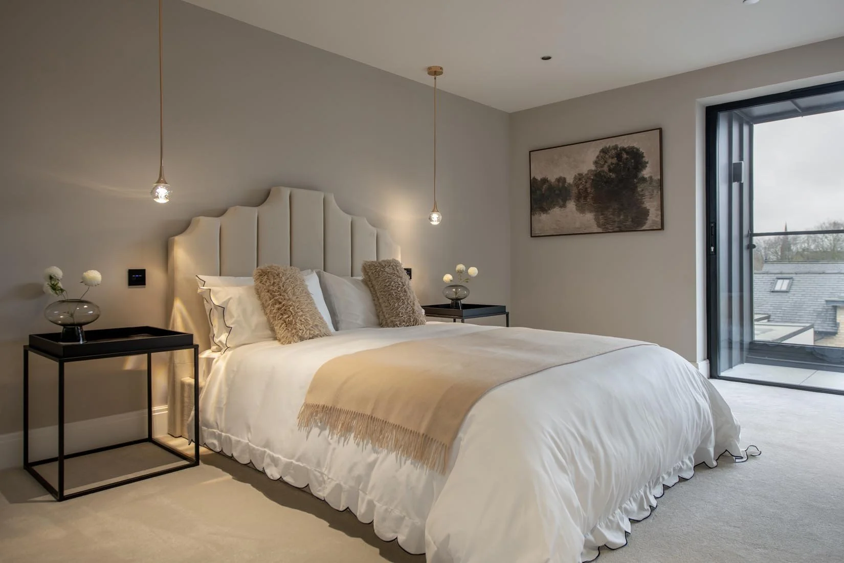 Modern bedroom with a large bed, beige headboard, white bedding, and beige throw blanket. Two black nightstands with glass vases holding white flowers. Two pendant lights hanging from the ceiling, a framed landscape painting, and a window with a balc