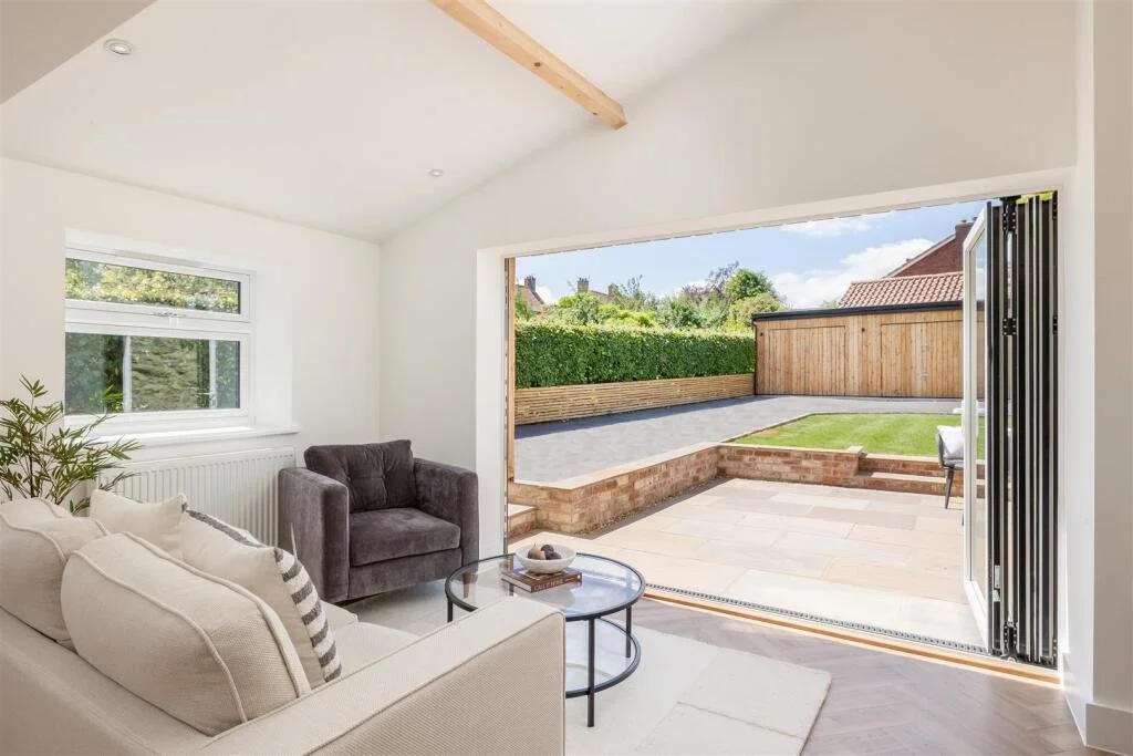 Interior living room with white walls, beige couch, dark gray armchair, coffee table, and a window, opening to a backyard with a patio, lawn, and wooden fence.