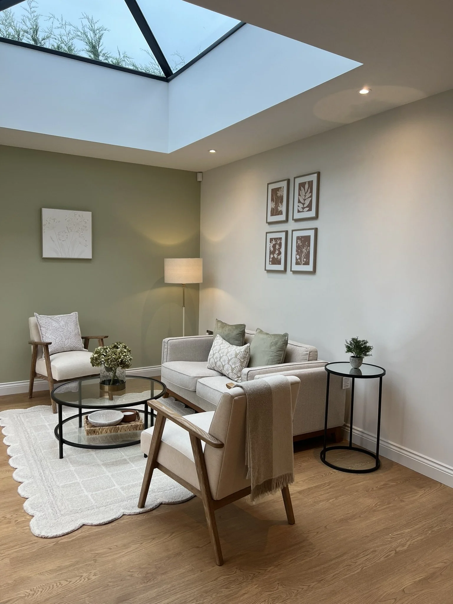 Living room with beige sofa, armchair, glass coffee table, side tables, framed botanical art, and a skylight.
