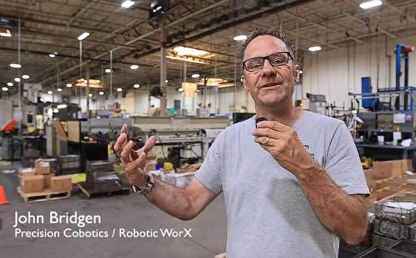 A man in a warehouse, wearing glasses and a gray t-shirt, gesturing with his hands, with industrial equipment and boxes in the background.