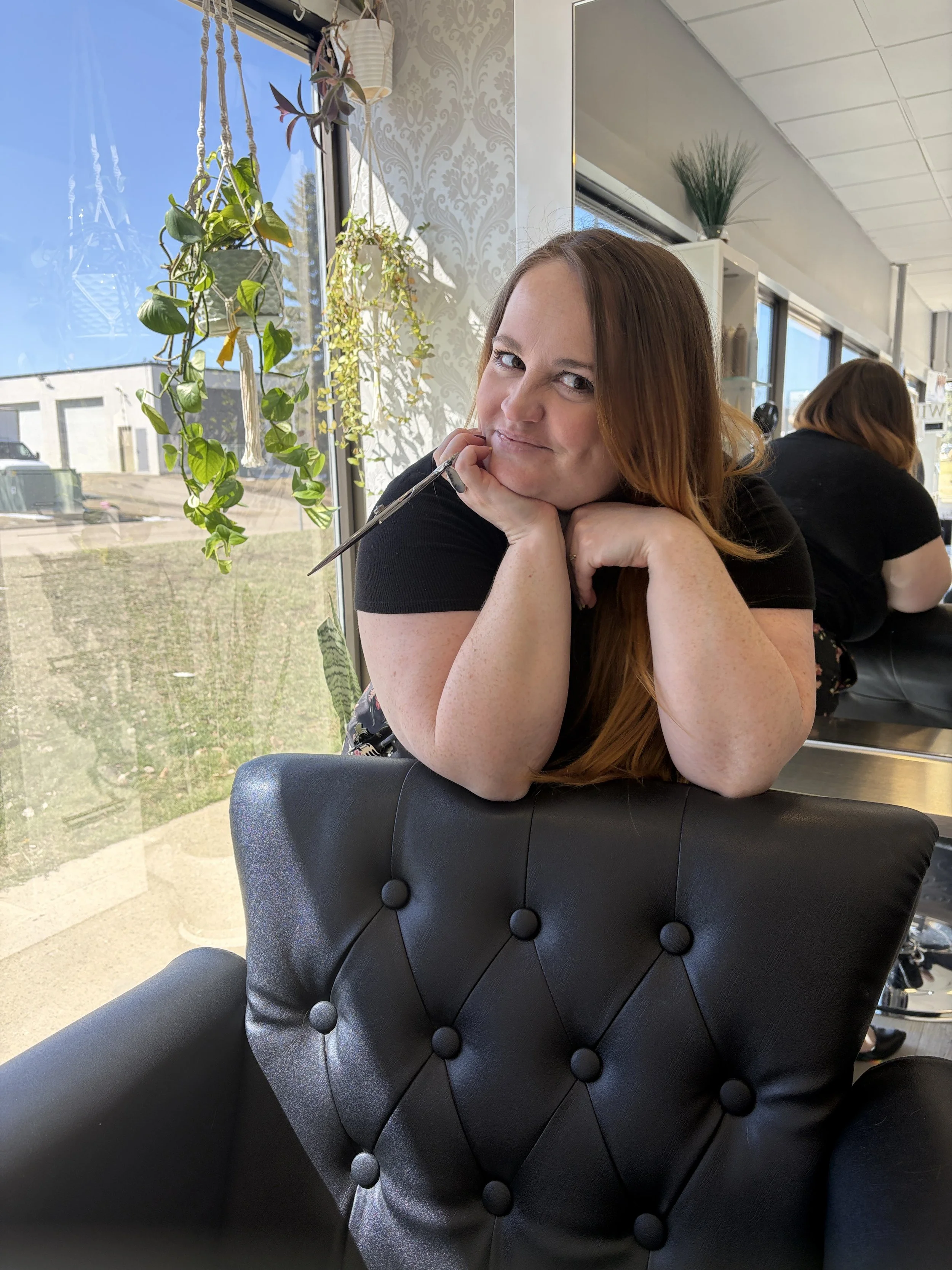 A woman with red hair sitting indoors at a table, resting her chin on her hands and smiling, with a window and hanging plants in the background.