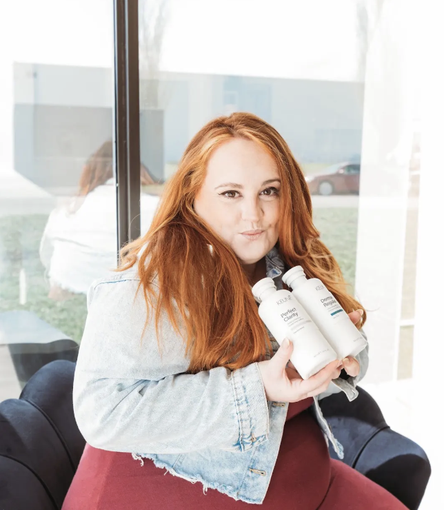 A woman with long red hair, wearing a black shirt, is seated at a table with a black, quilted leather chair in front of her. She is resting her chin on her hands and smiling at the camera inside a brightly lit cafe or restaurant. The background includes a window with hanging plants, patterned wallpaper, and another person with similar hair color facing away.