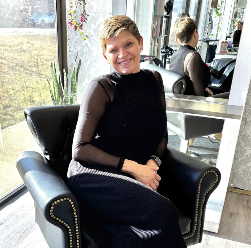 A woman with short hair, smiling, sitting in a black leather chair in a salon, with a mirror behind her reflecting her back and the salon interior.