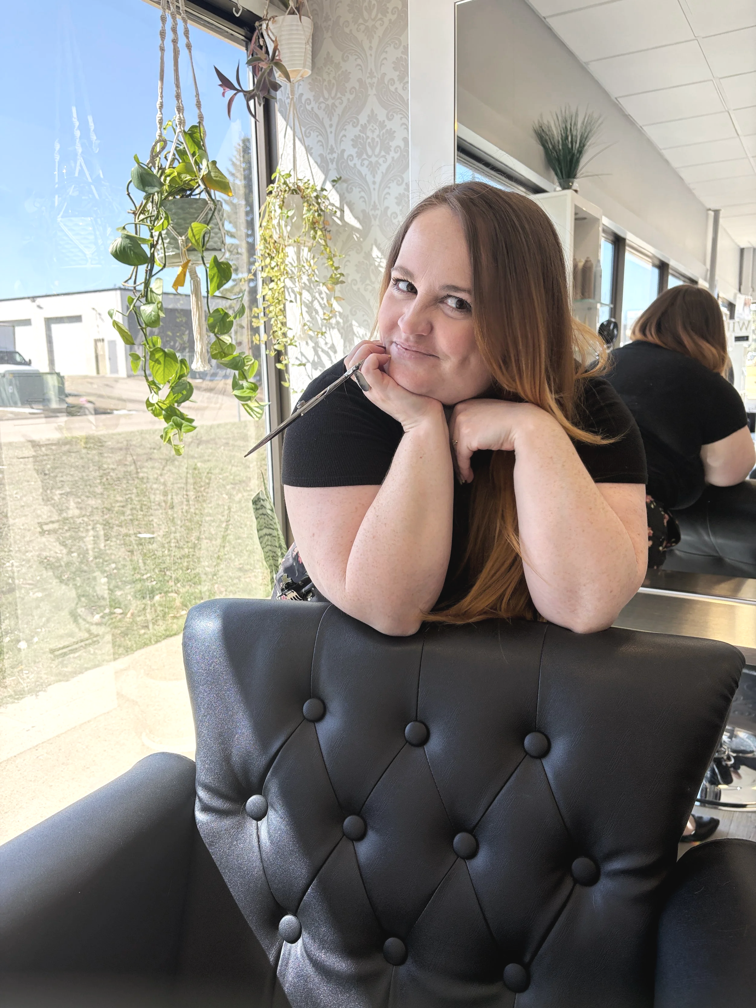 A woman with long red hair, wearing a black shirt, is seated at a table with a black, quilted leather chair in front of her. She is resting her chin on her hands and smiling at the camera inside a brightly lit cafe or restaurant. The background includes a window with hanging plants, patterned wallpaper, and another person with similar hair color facing away.