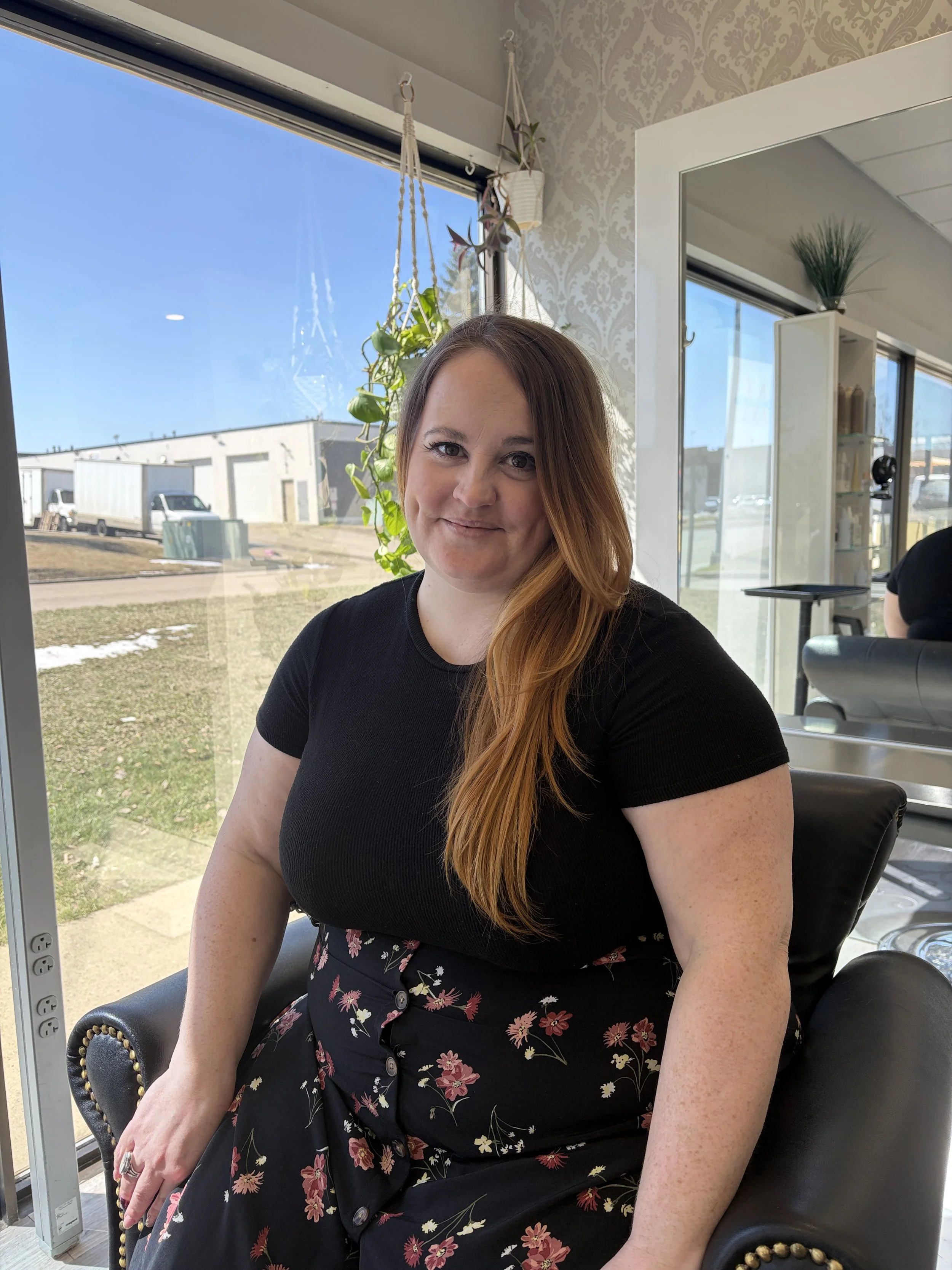 A woman with long red hair and fair skin, wearing a black top and a black skirt with pink and white floral print, sitting in a black armchair with gold nailhead trim near a large window with a view of a sunny outdoor scene. She is smiling softly at the camera inside a well-lit room with patterned wallpaper, hanging plants, and decorative items.