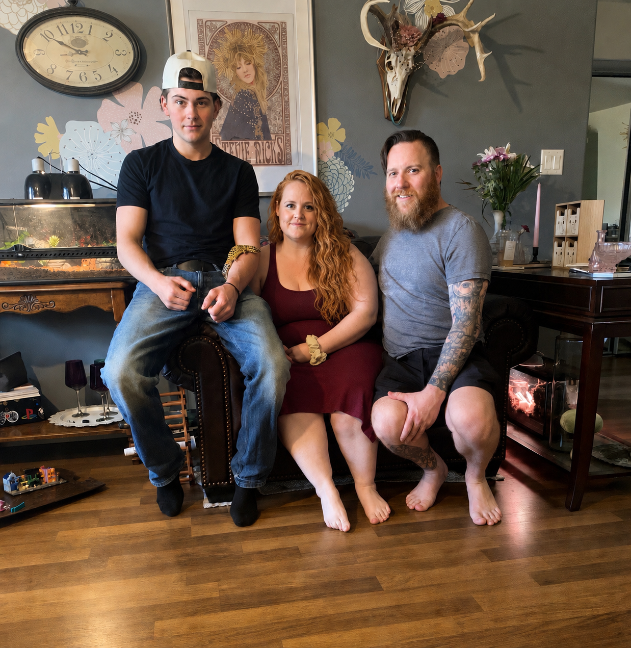A family of three sitting and standing on a dark brown leather couch in a decorated living room. The young man on the left is wearing a black t-shirt, jeans, and a backwards white cap, with a yellow and black snake on his arm. The woman in the middle has long curly red hair and is dressed in a sleeveless maroon dress. The man on the right has a full beard, tattoos on his left arm, and is wearing a gray t-shirt and black shorts. The background features a decorative wall with artwork, a mounted deer skull, and a clock. To their left, there's an aquarium with lighting and decor, and on the right, a wooden table with flowers and decorative items.