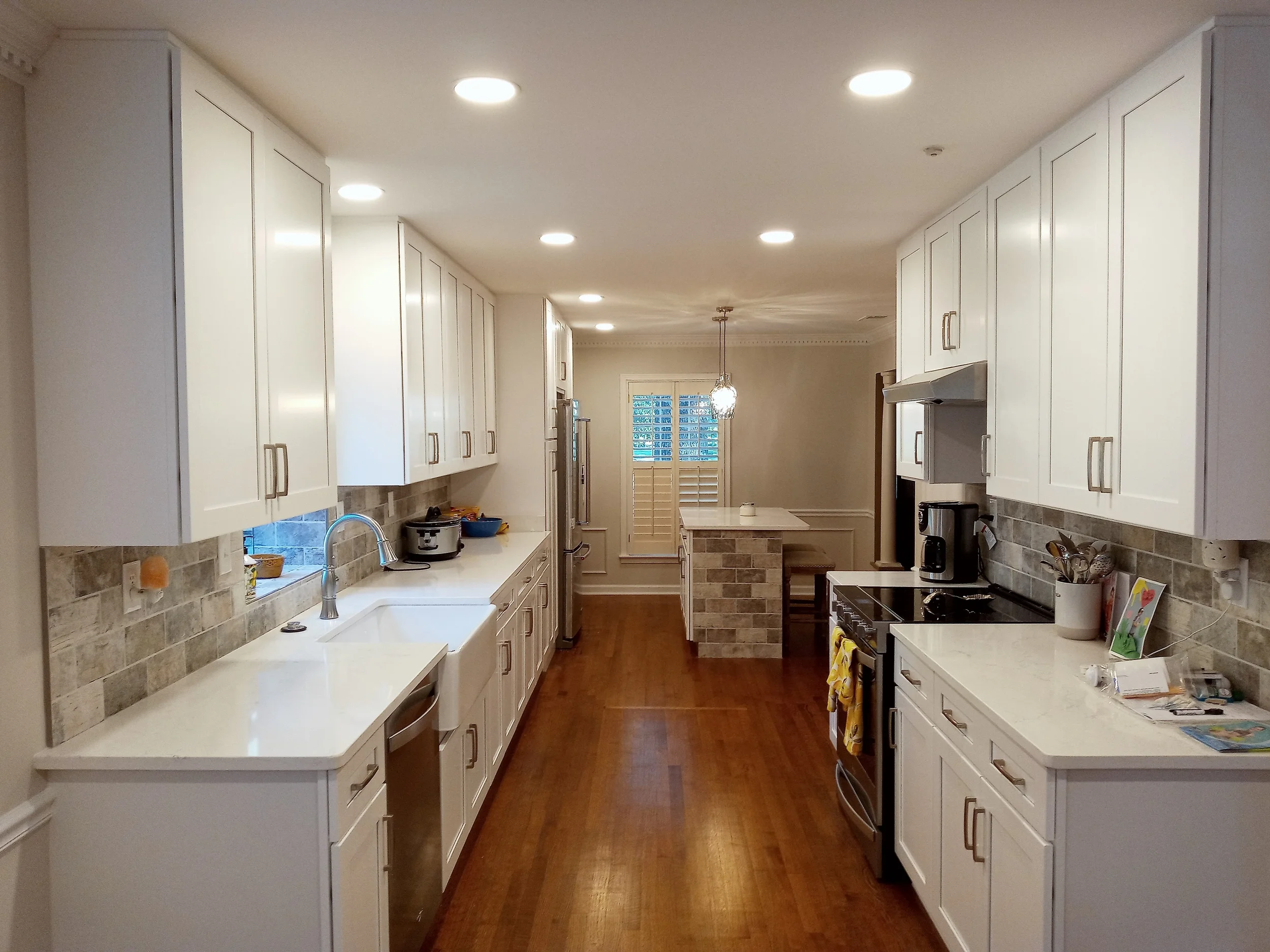 A modern kitchen with white cabinets, a white countertop, and a brick backsplash, featuring a sink, stove, and small kitchen island, with hardwood floors and recessed lighting.