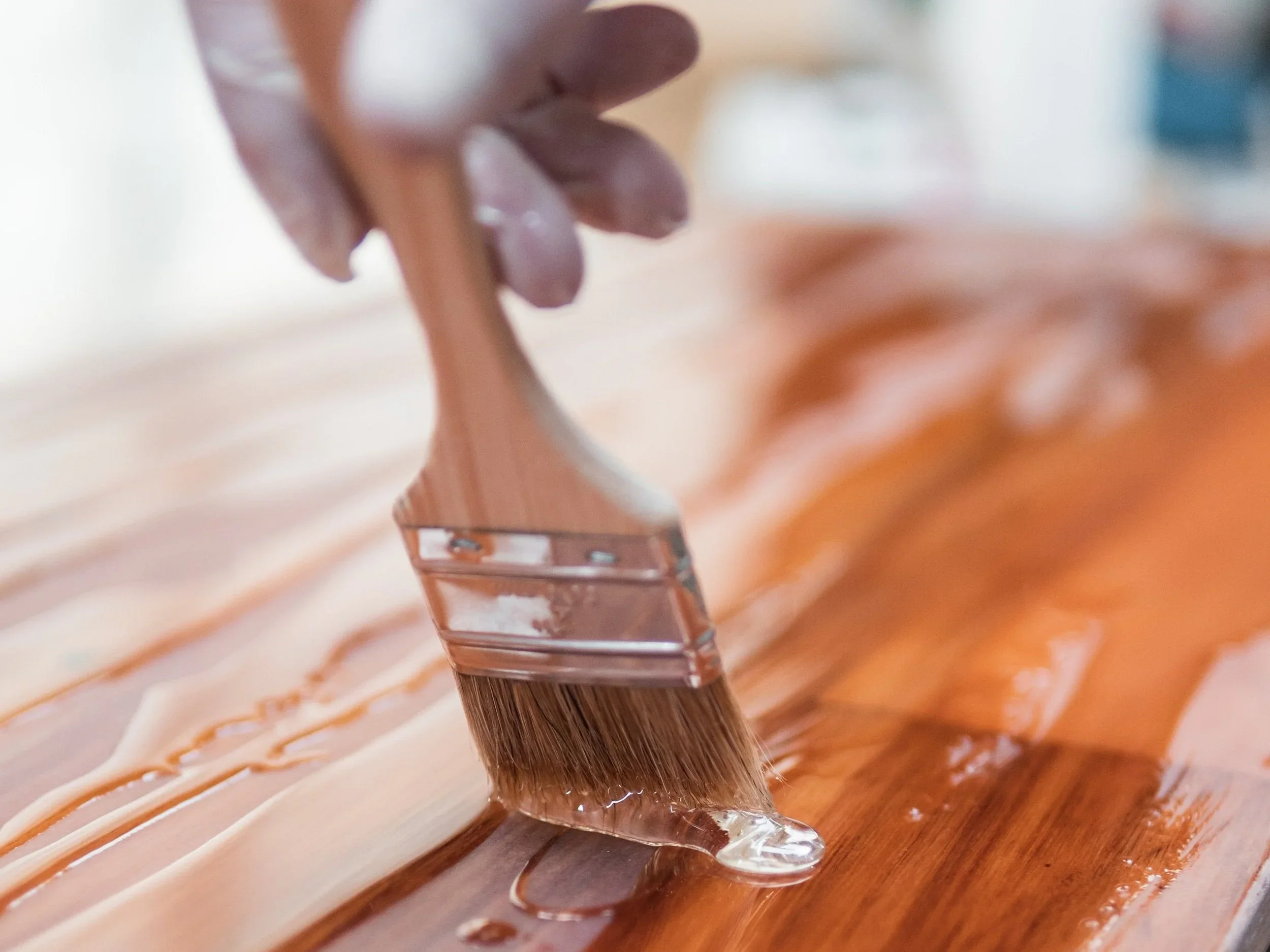 Close-up of a hand holding a paintbrush, applying varnish to a wooden surface.