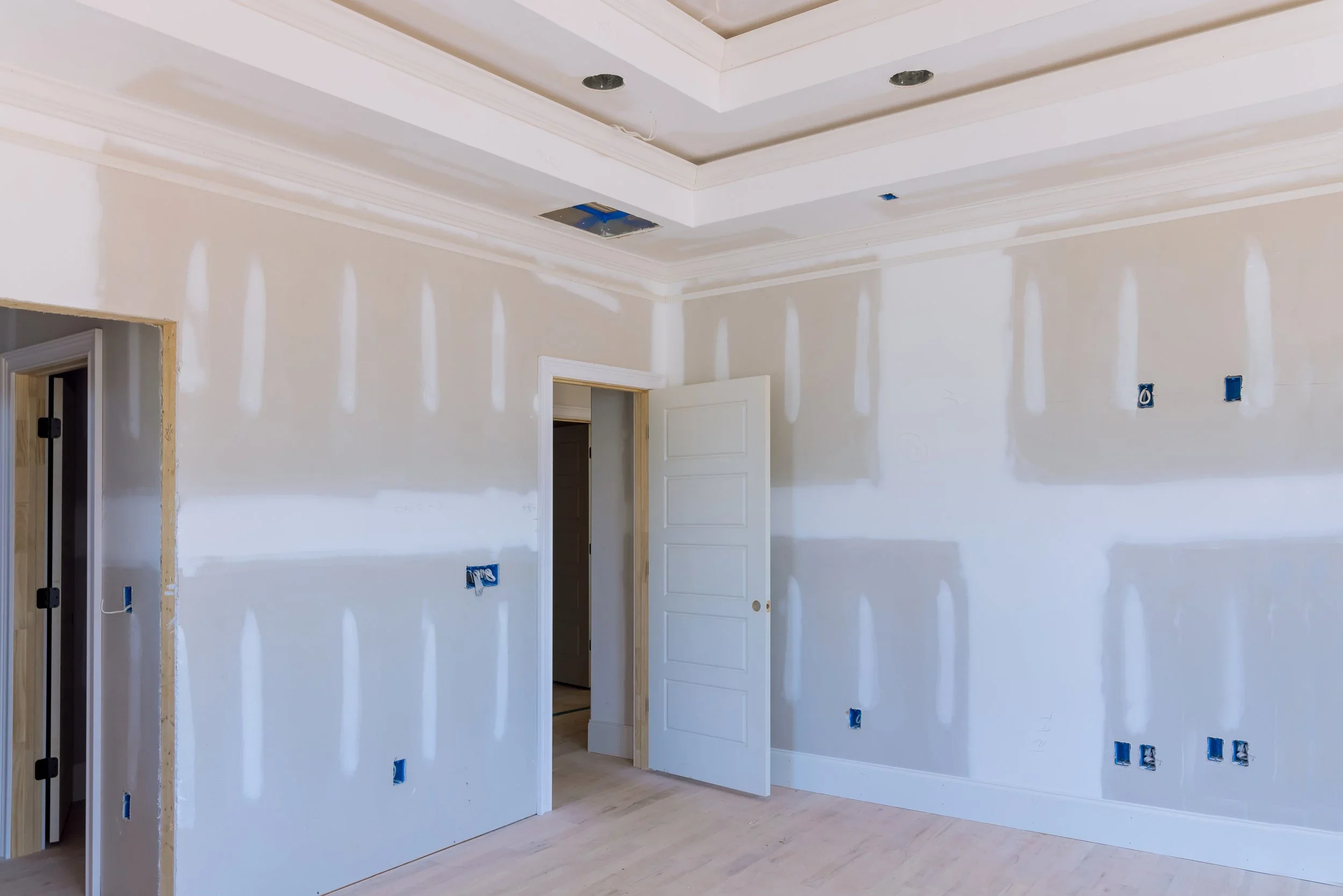 An unfinished room with drywall installed, light-colored wooden flooring, and open electrical outlets and switches. The ceiling has decorative molding with some cutouts or fixtures missing.